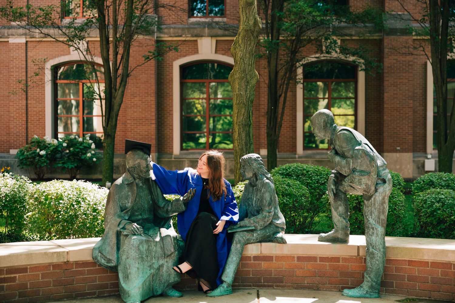 Graduate in blue academic regalia sits among sculptural figures on campus brick wall.