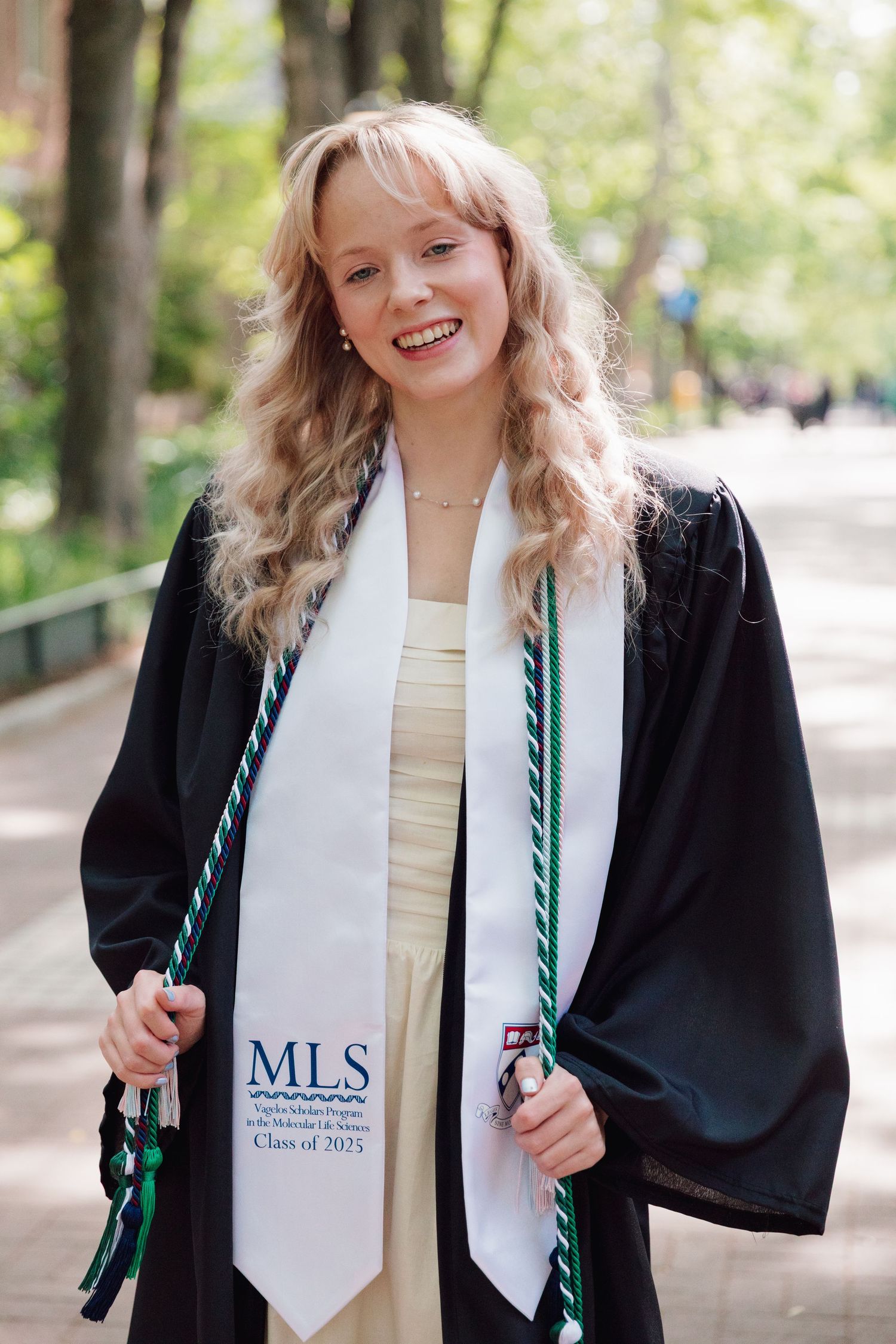 Graduate wearing MLS sash and graduation gown smiles outdoors on campus pathway.