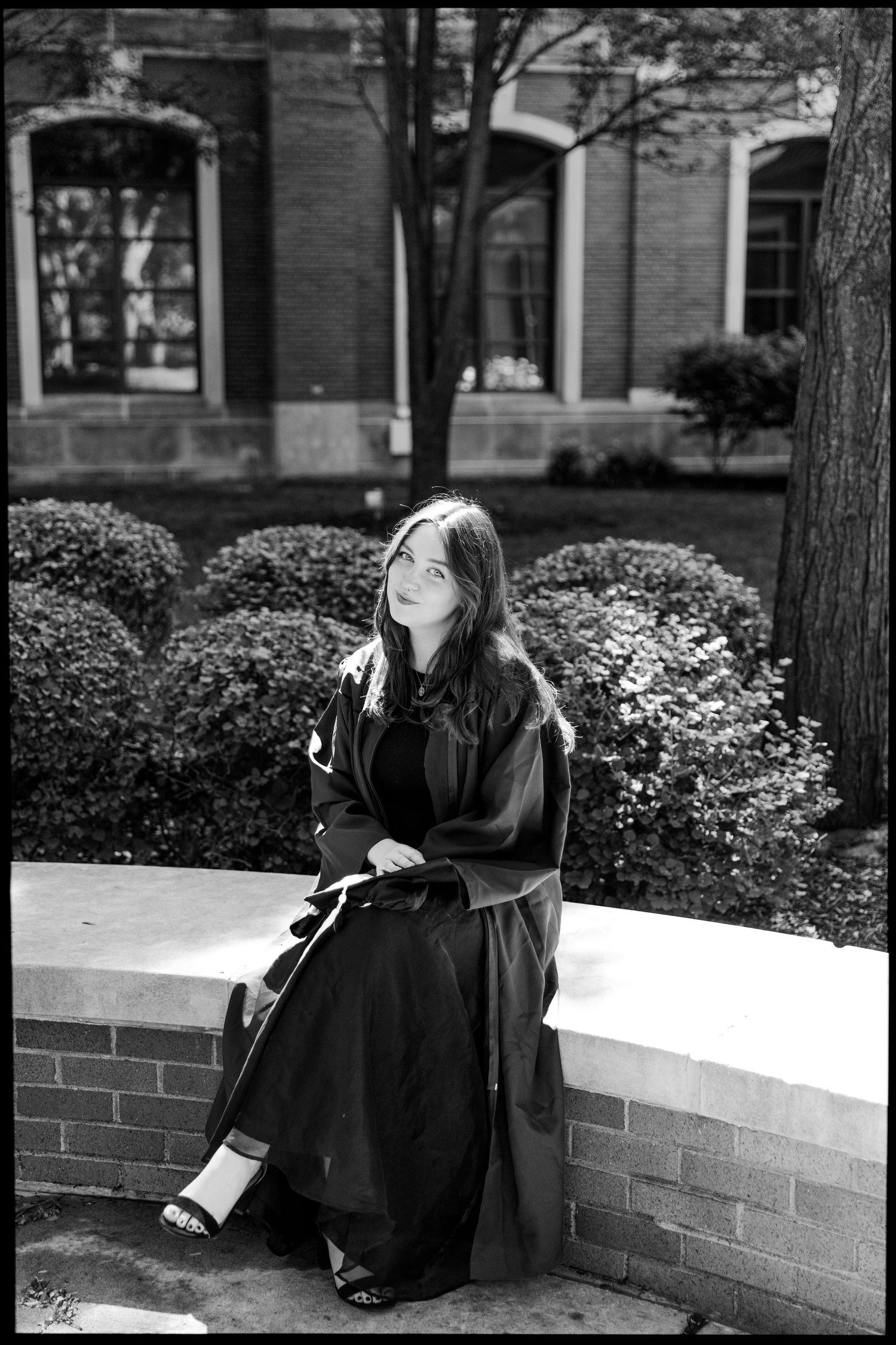 Black and white portrait of graduate sitting on brick wall in academic regalia.