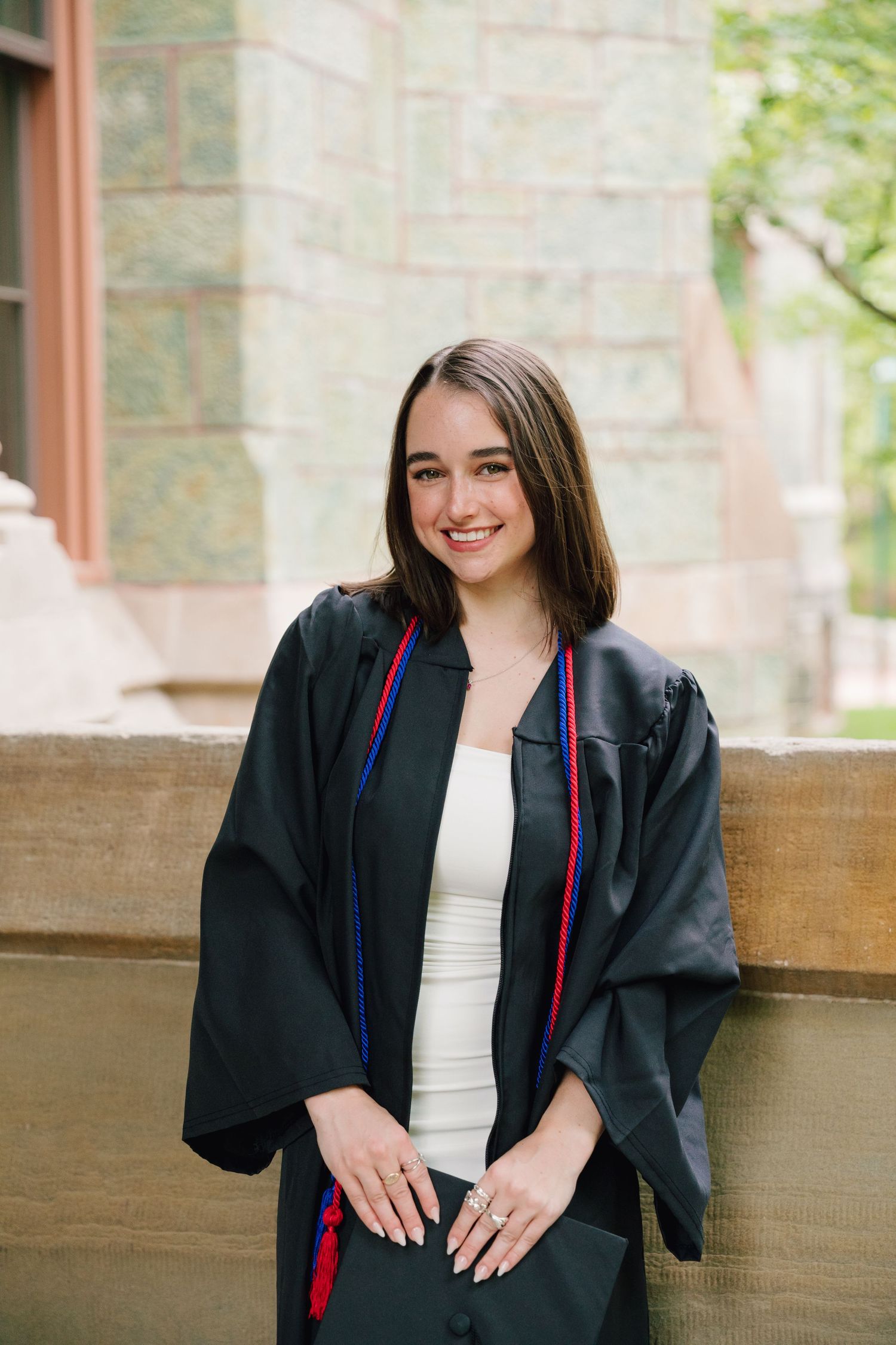 Graduate in black gown poses against stone building wall.