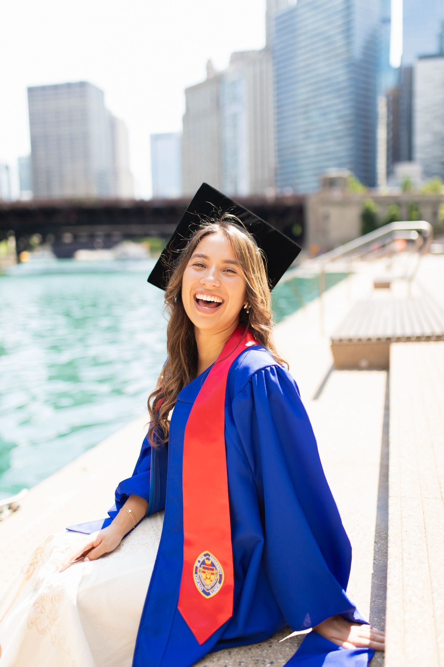 Graduate in blue and red regalia poses joyfully along Chicago River with downtown skyline in background.