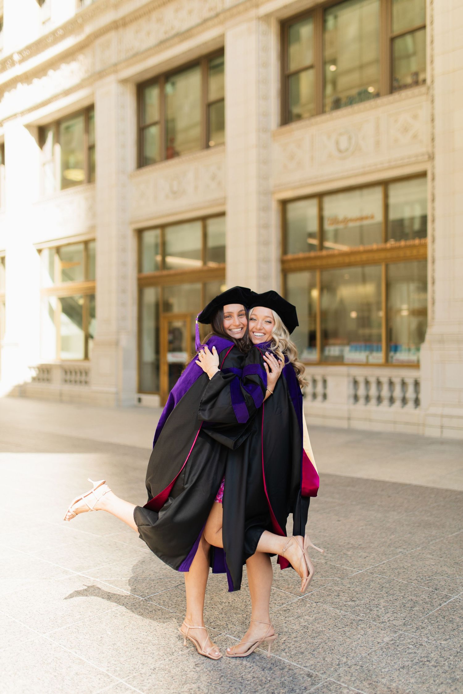 Two graduates in purple academic regalia embrace and pose together outdoors.