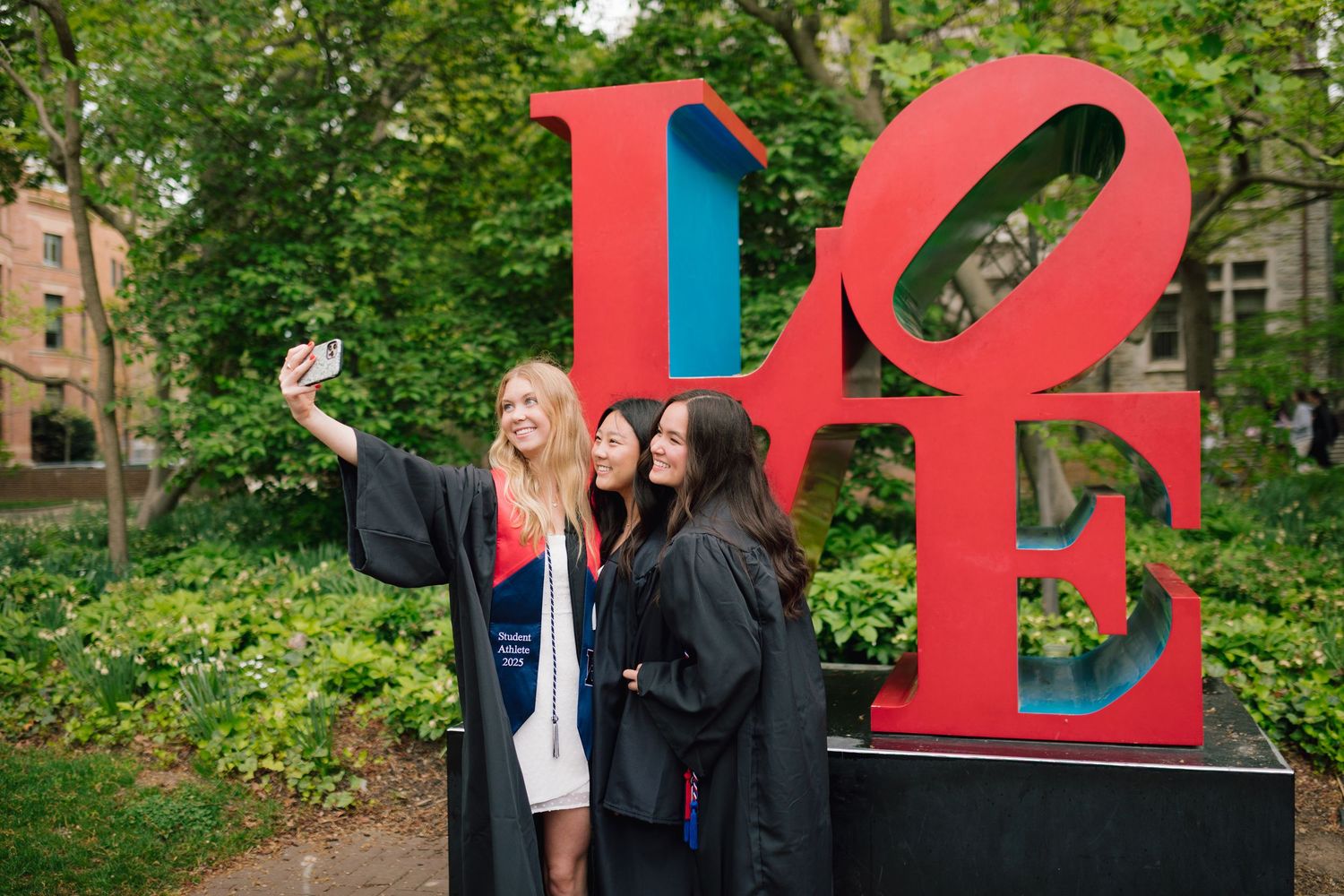 Graduates pose for photos with large red LOVE sculpture on university campus.