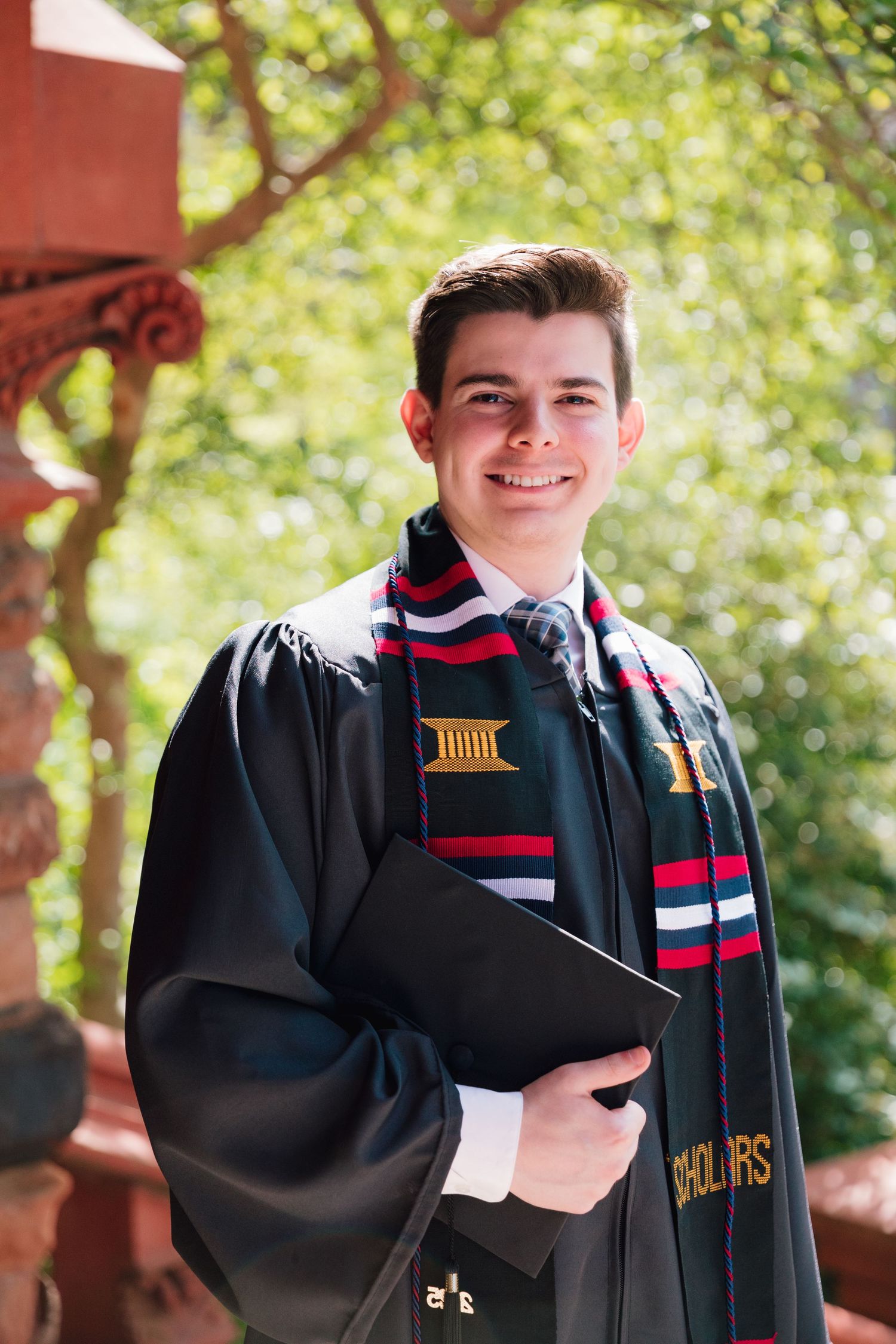 Graduate in black academic regalia poses with colorful stole against red brick architecture.