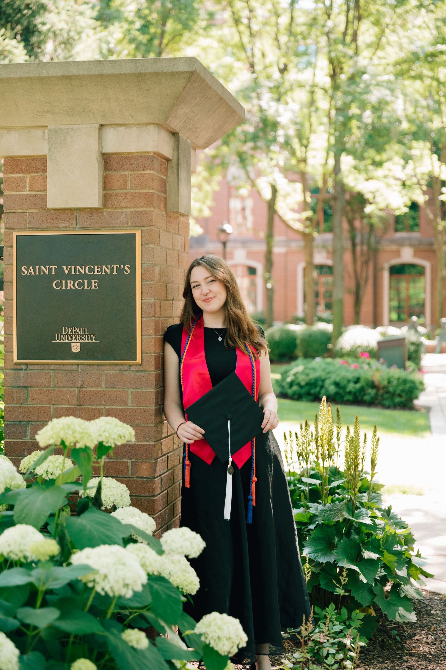 Graduate in academic regalia poses by campus signage surrounded by white hydrangea bushes.