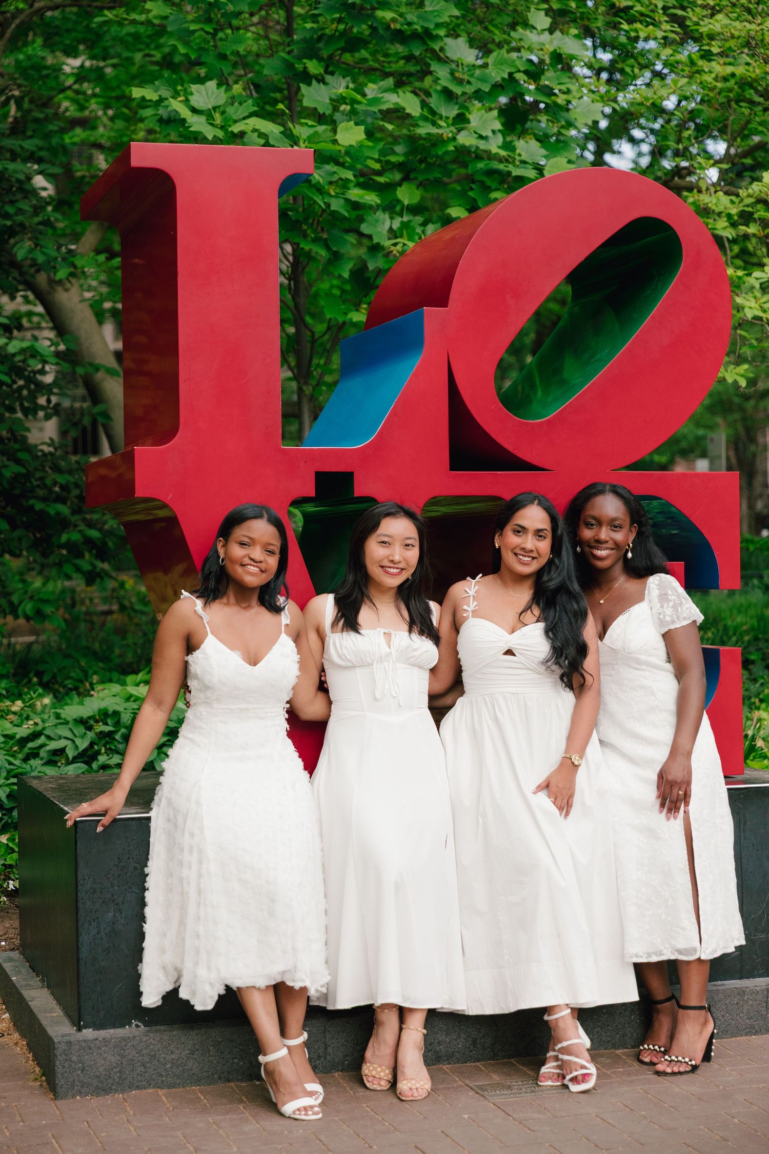 Graduates in white dresses pose together in front of red LOVE sculpture.