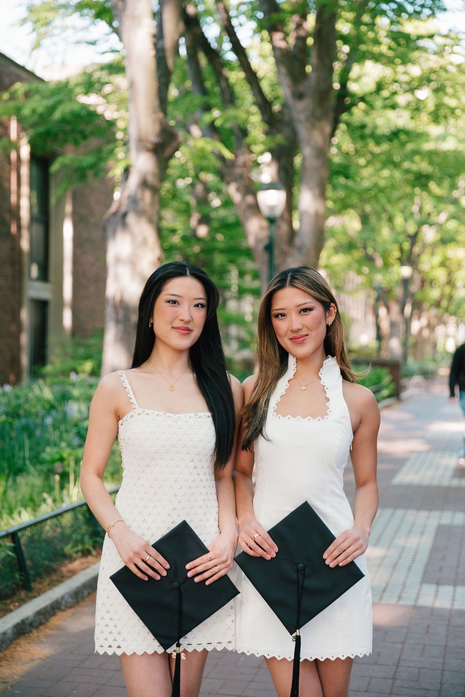 Two graduates in white dresses hold their graduation caps on a tree-lined university walkway.