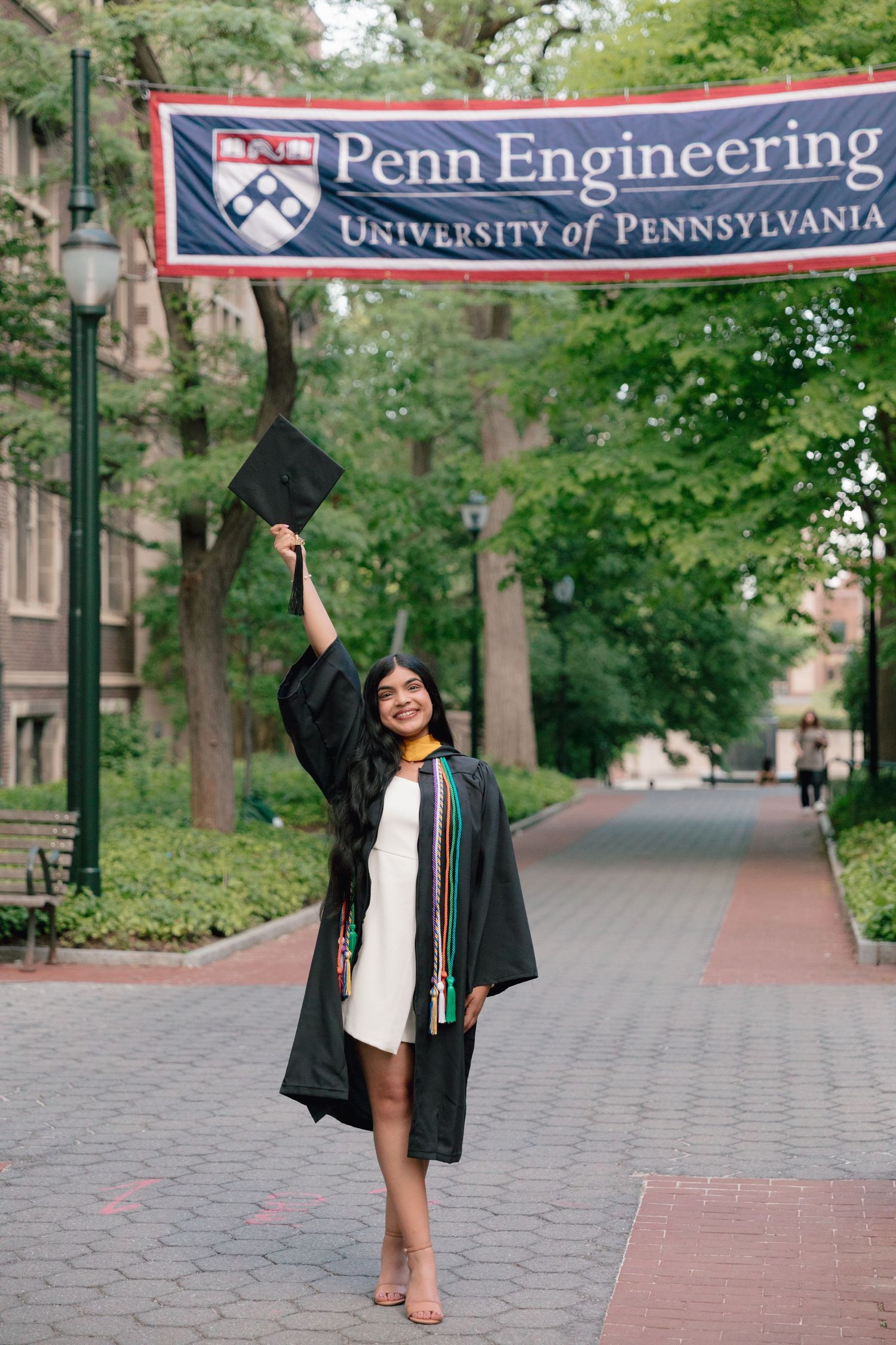 Graduate celebrating under Penn Engineering banner on campus walkway.