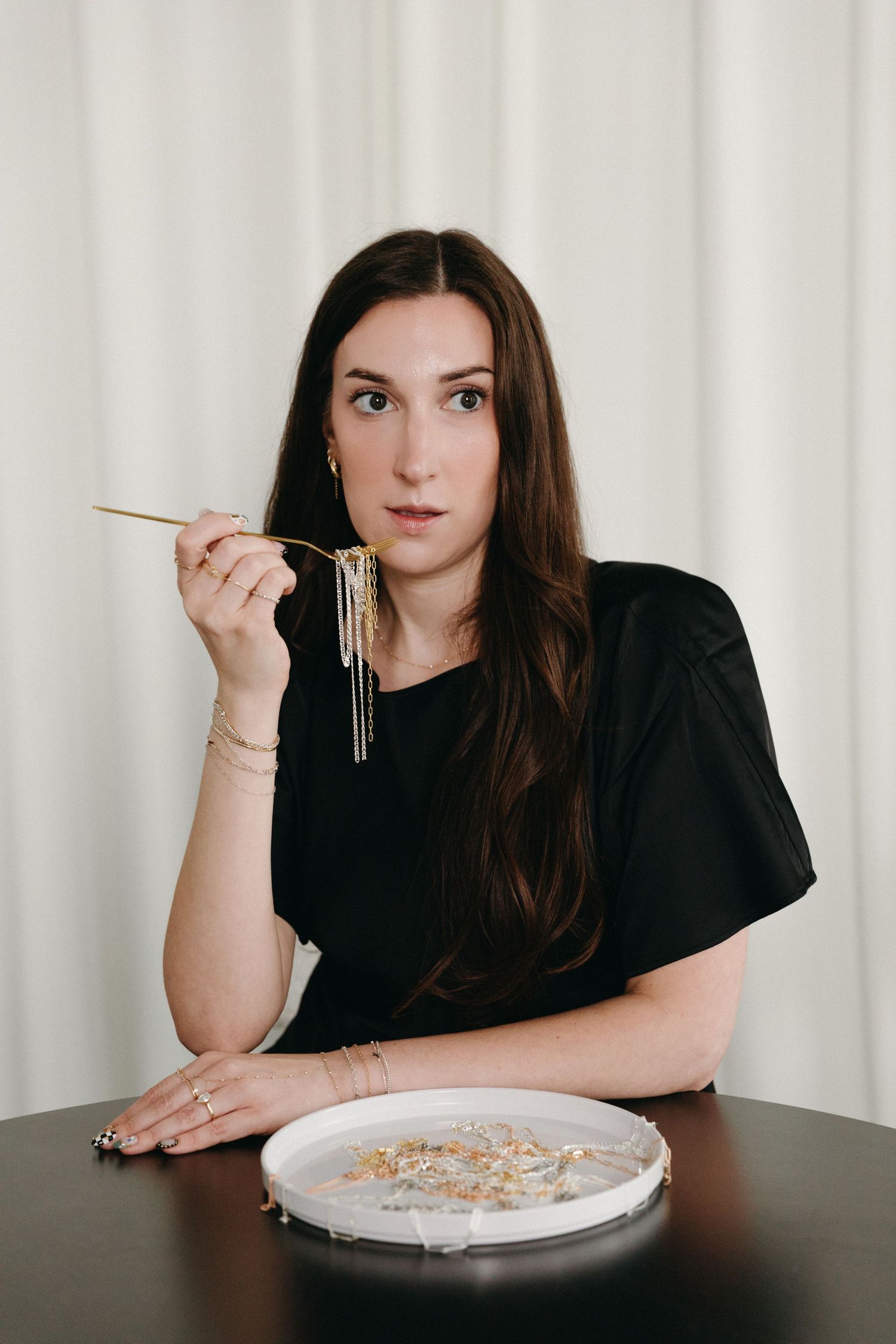Woman in black top eating noodles with chopsticks at a dark table, white curtain background.