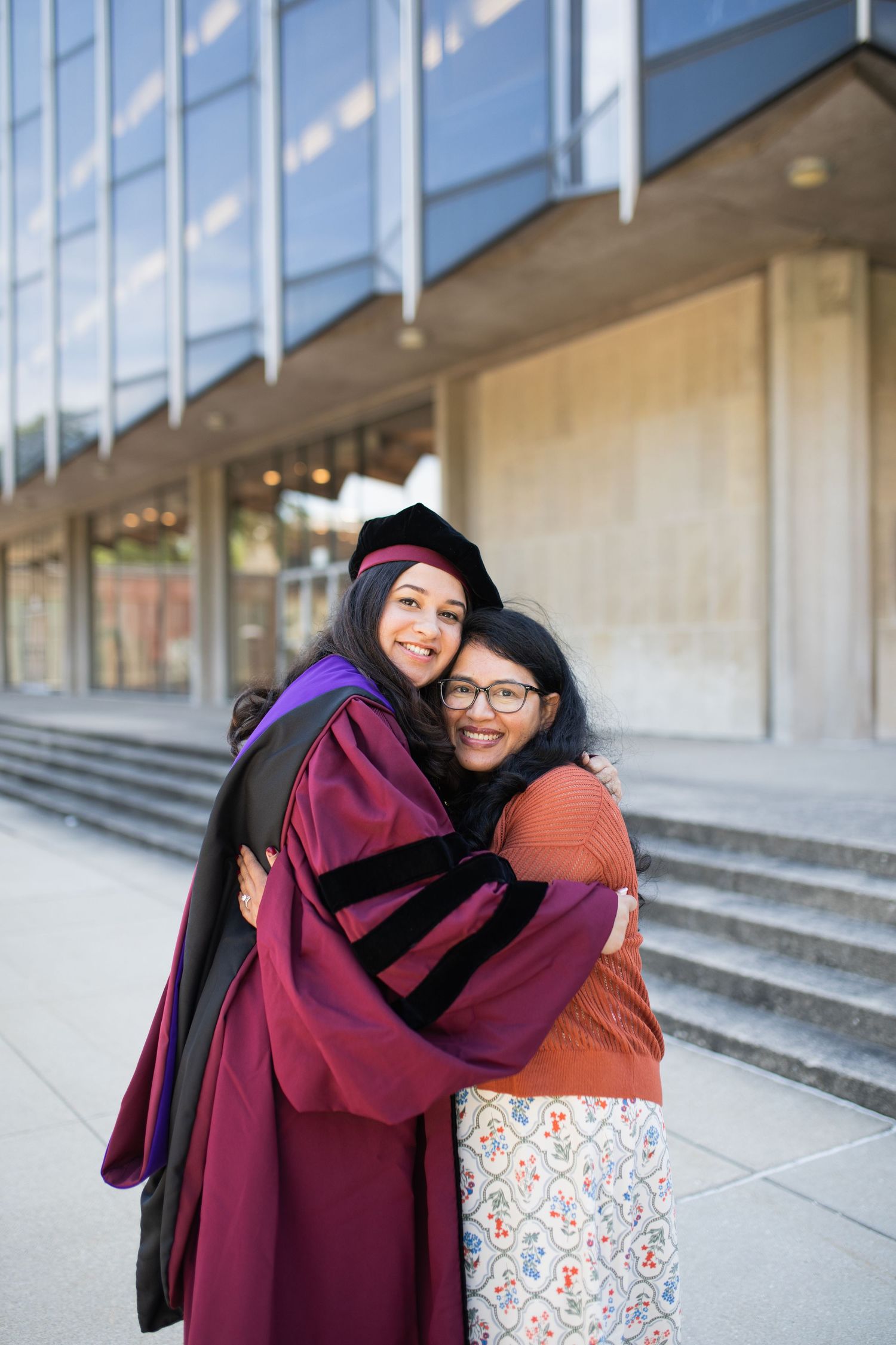 Two graduates embrace in celebration outside modern building entrance.