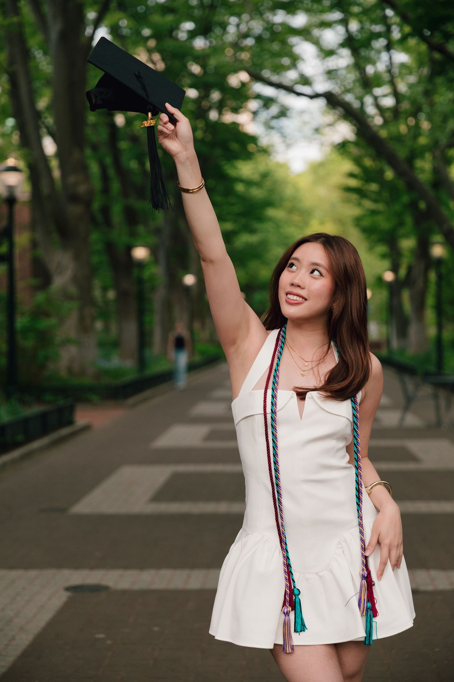 Graduate in white dress tosses cap in celebration on tree-lined university path.