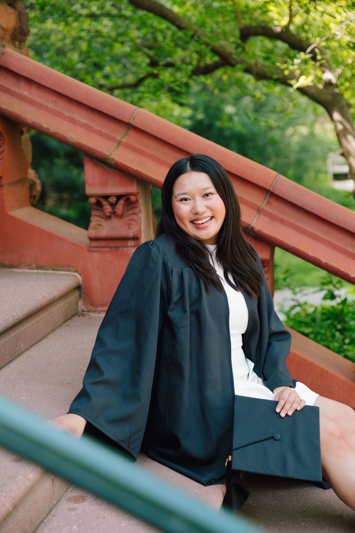 Graduate in academic regalia poses on red stone steps surrounded by greenery.