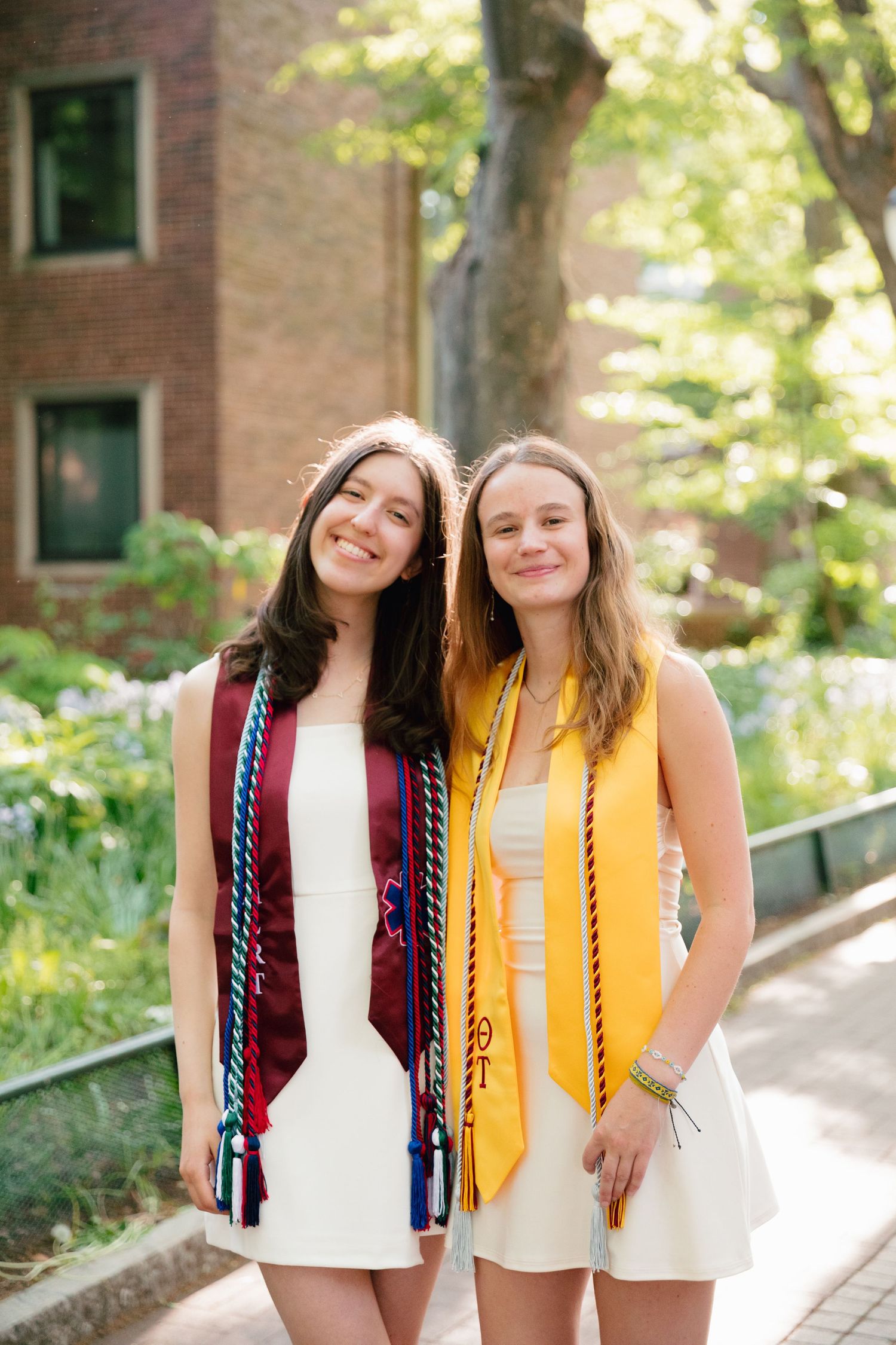 Two graduates in white dresses with honor cords pose on sunny campus path.