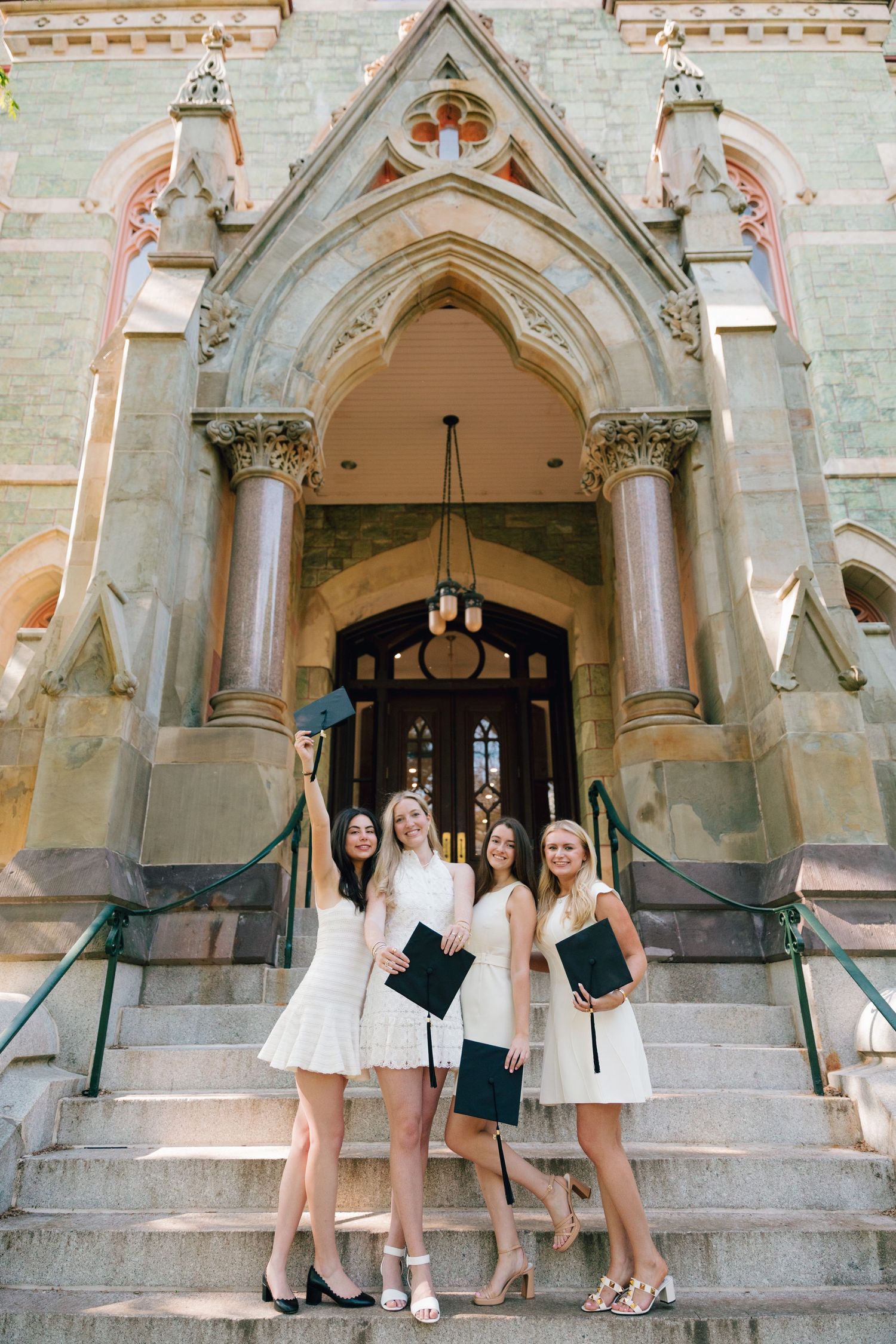 Four graduates in white dresses pose together on church steps under Gothic architecture.