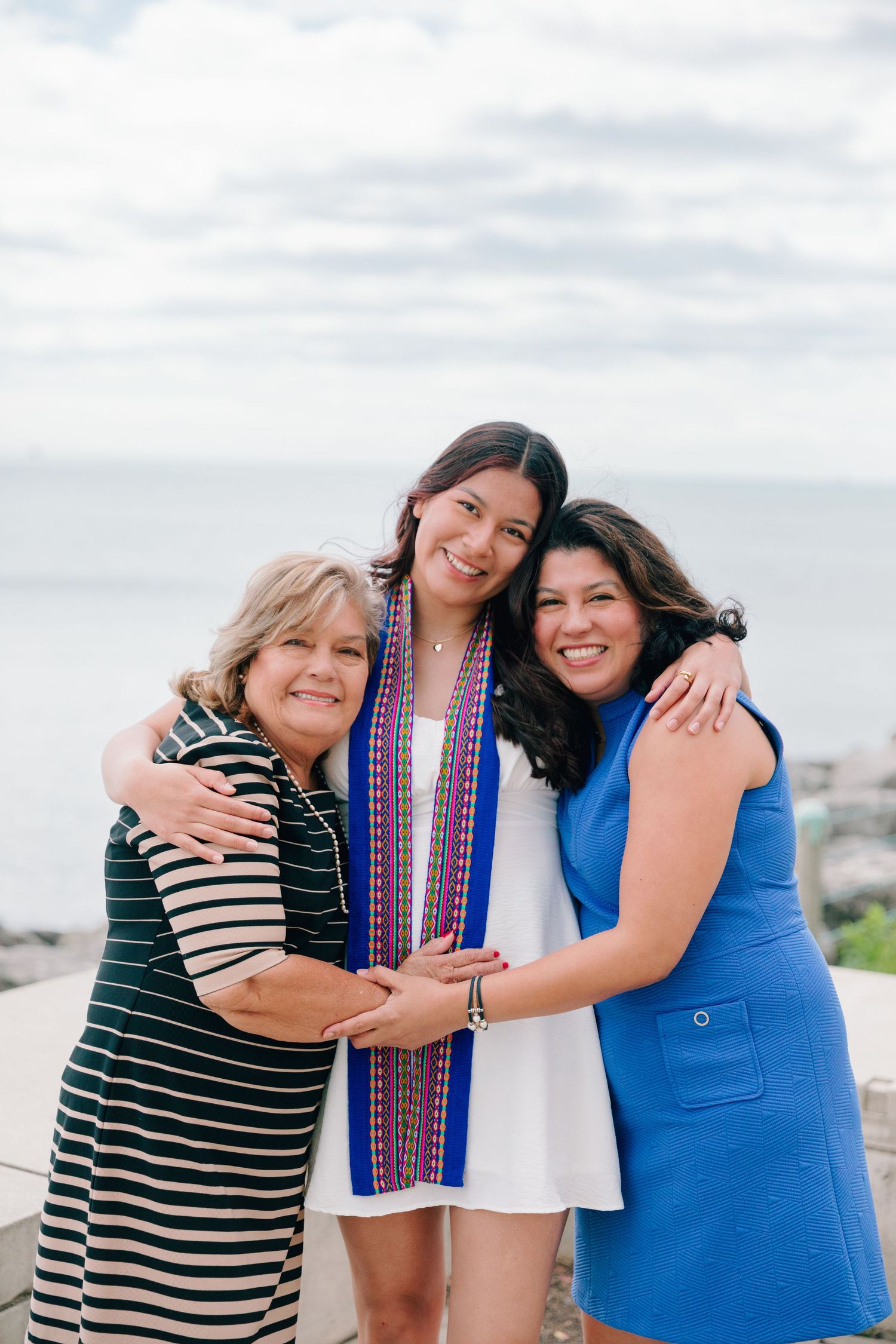 Three people embrace while celebrating graduation outdoors against cloudy sky.