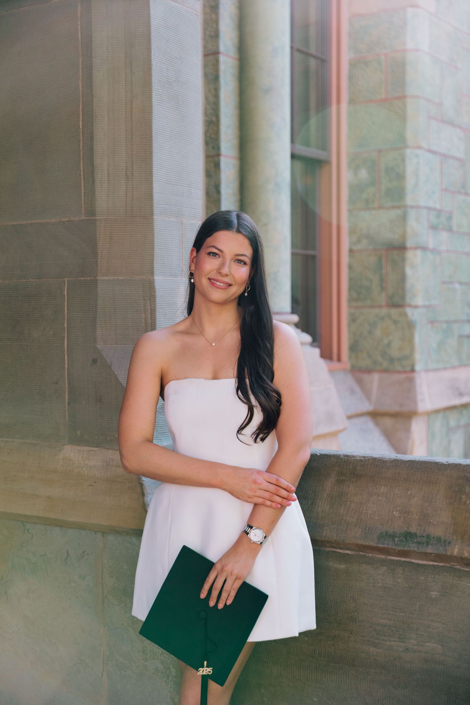Graduate in white top poses with diploma against stone column backdrop.