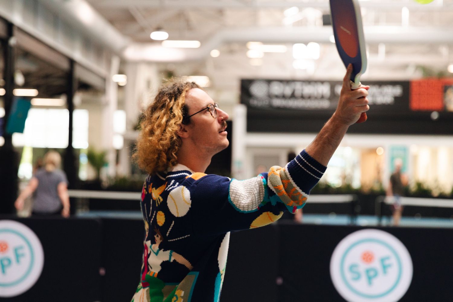 Woman in colorful dress playing pickleball indoors, raising paddle overhead at SPF tournament event.