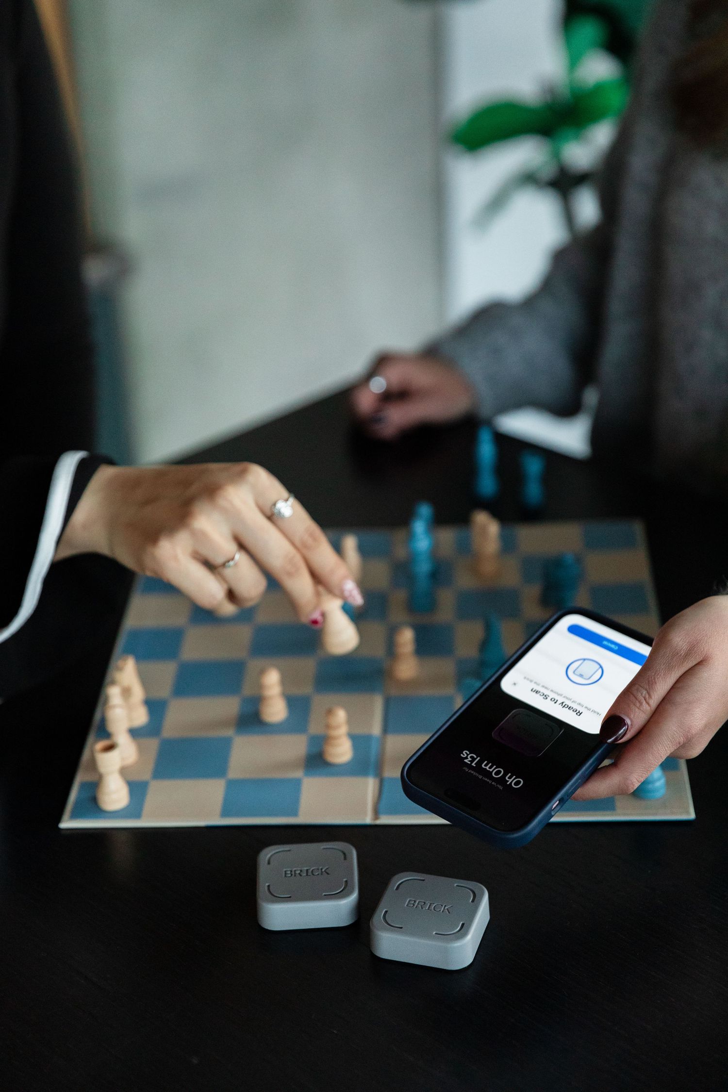 Two people playing chess with a smartphone displaying an app and small devices on a dark table.