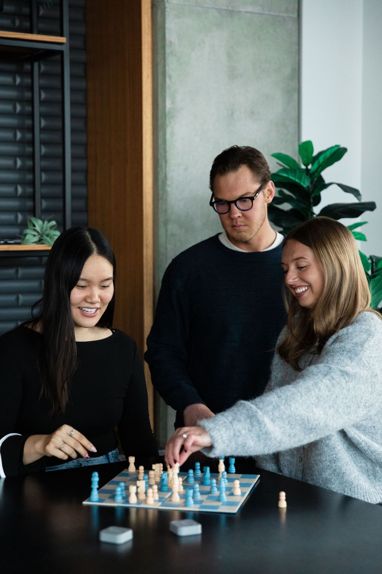 Three people smiling and playing a blue and white board game together at a dark table in a modern office.