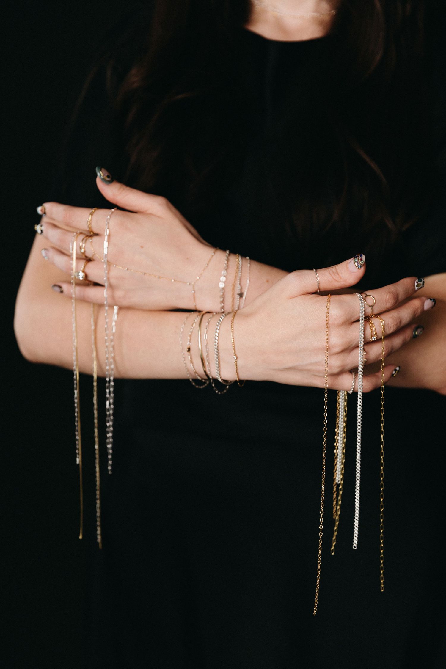 Woman displaying multiple gold chain bracelets and rings stacked on crossed hands against black background.