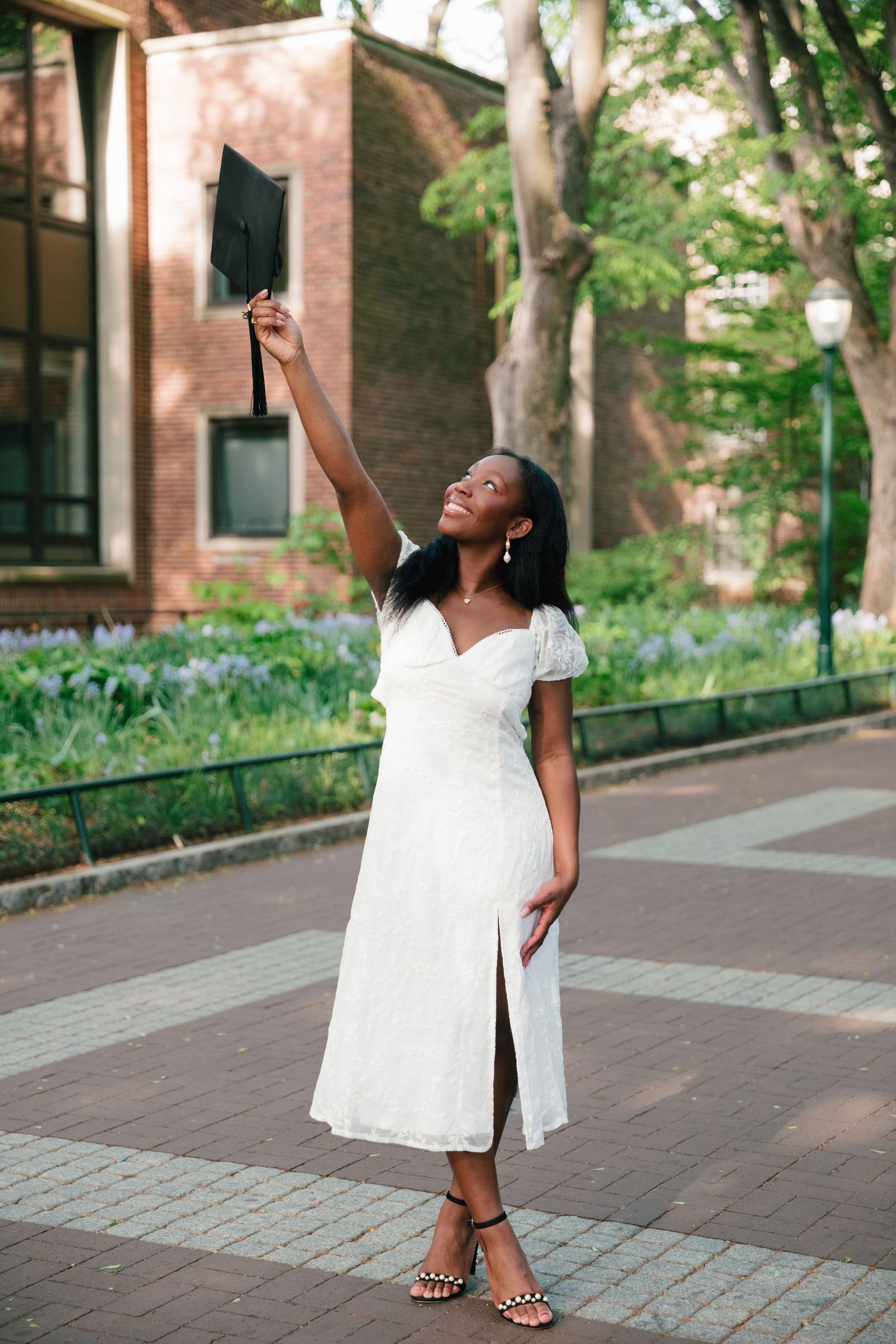 Graduate in white dress raises cap joyfully on campus walkway.