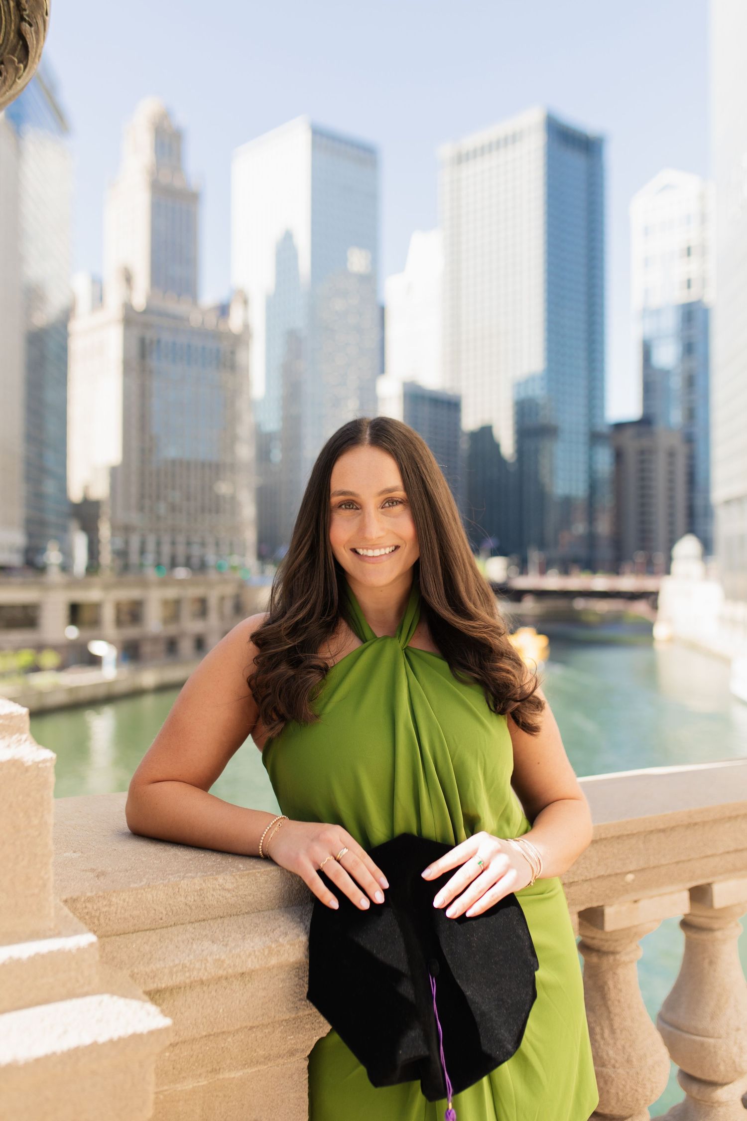 Graduate in green dress poses with diploma against Chicago skyline and river backdrop.