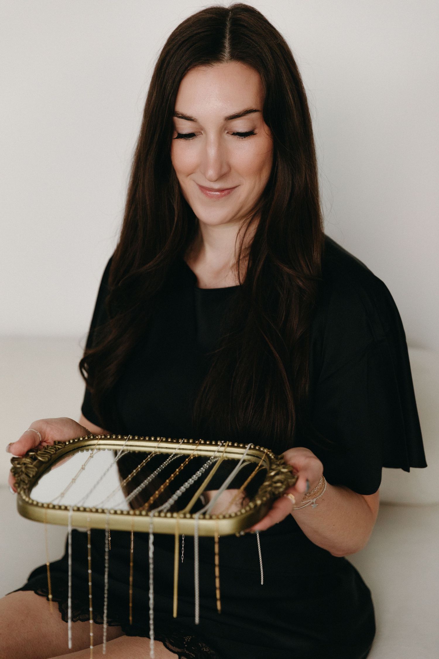 Woman in black dress holding a gold ornate tray with hanging necklaces, smiling softly in a bright room.