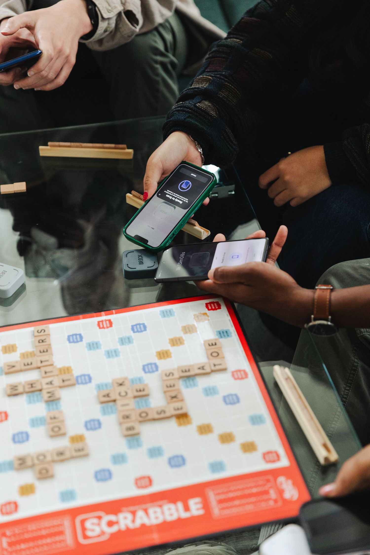 Friends playing Scrabble board game on glass table with tiles arranged, while one player uses a smartphone.