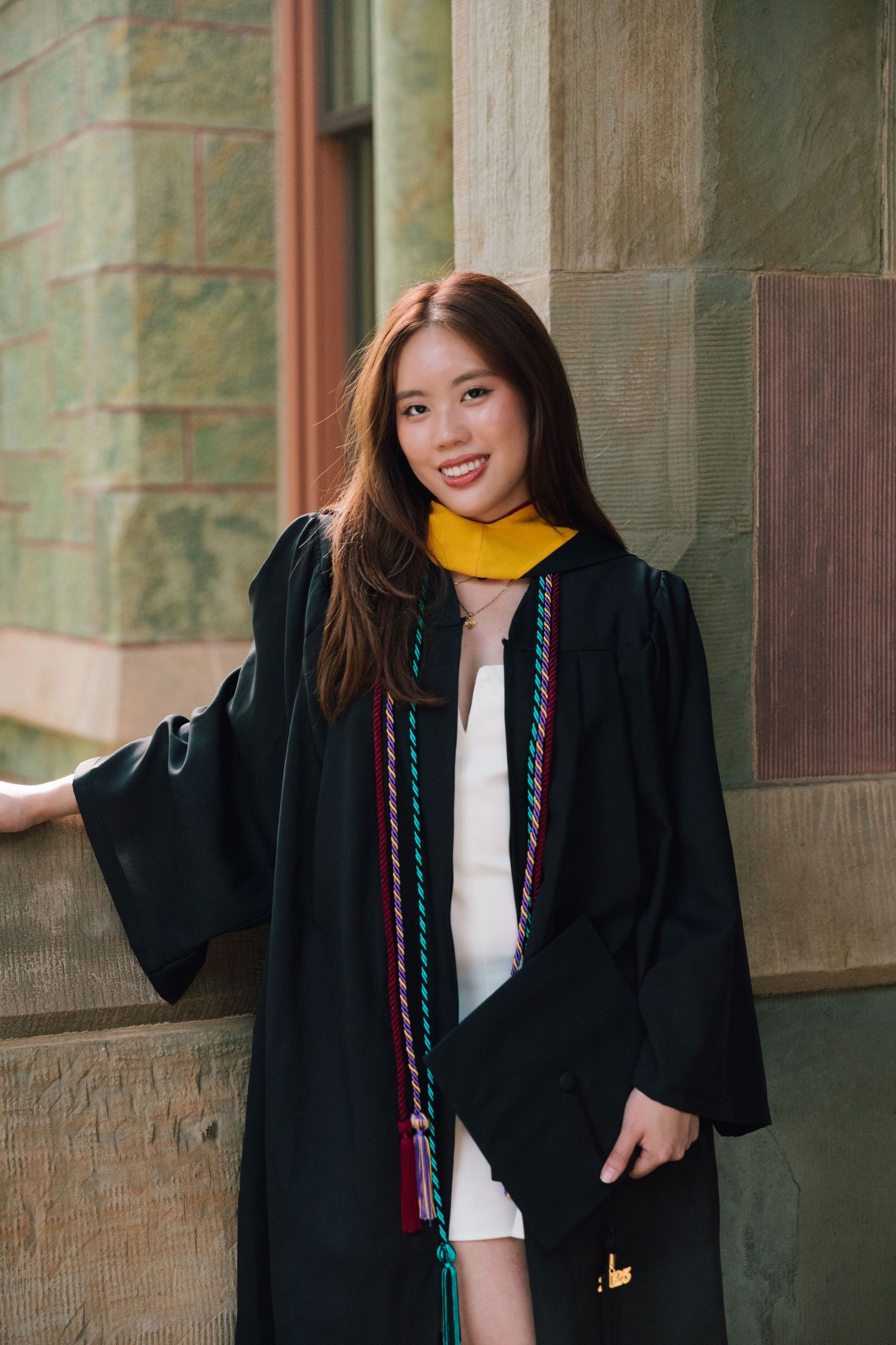Graduate in black cap and gown poses playfully against stone university building wall.