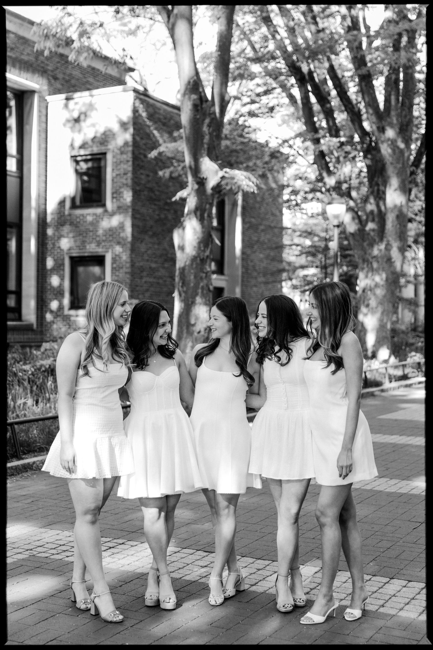 Group of graduates in white dresses pose together on brick pathway.