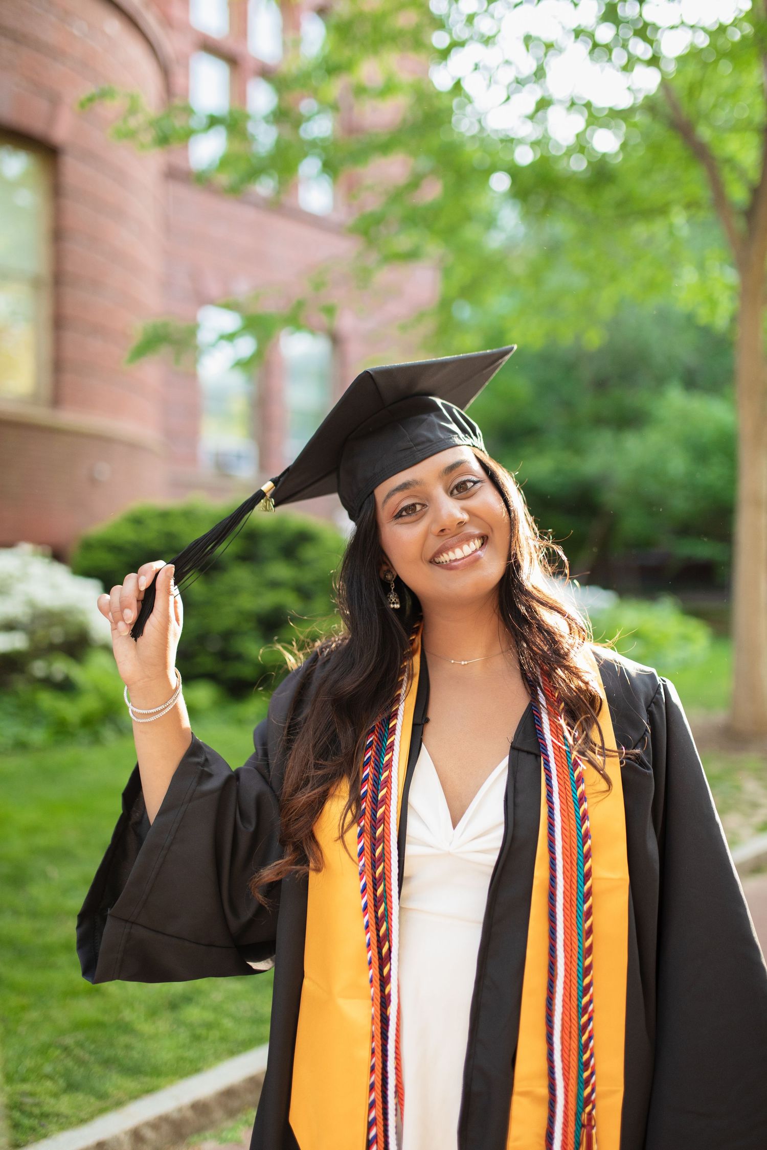 Graduate in black cap and gown with honor cords poses joyfully on campus grounds.