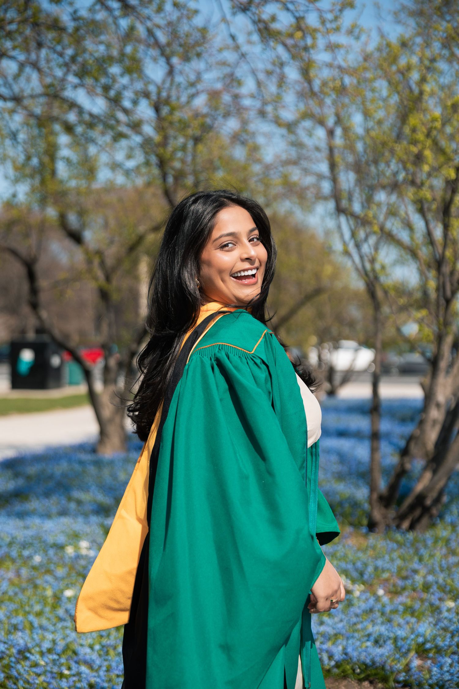 Graduate in emerald green gown poses joyfully among blooming blue flowers.