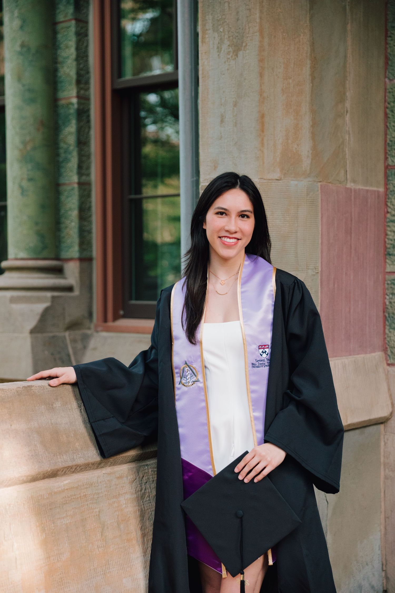 Graduate posing in cap and gown with purple stole against stone building wall.