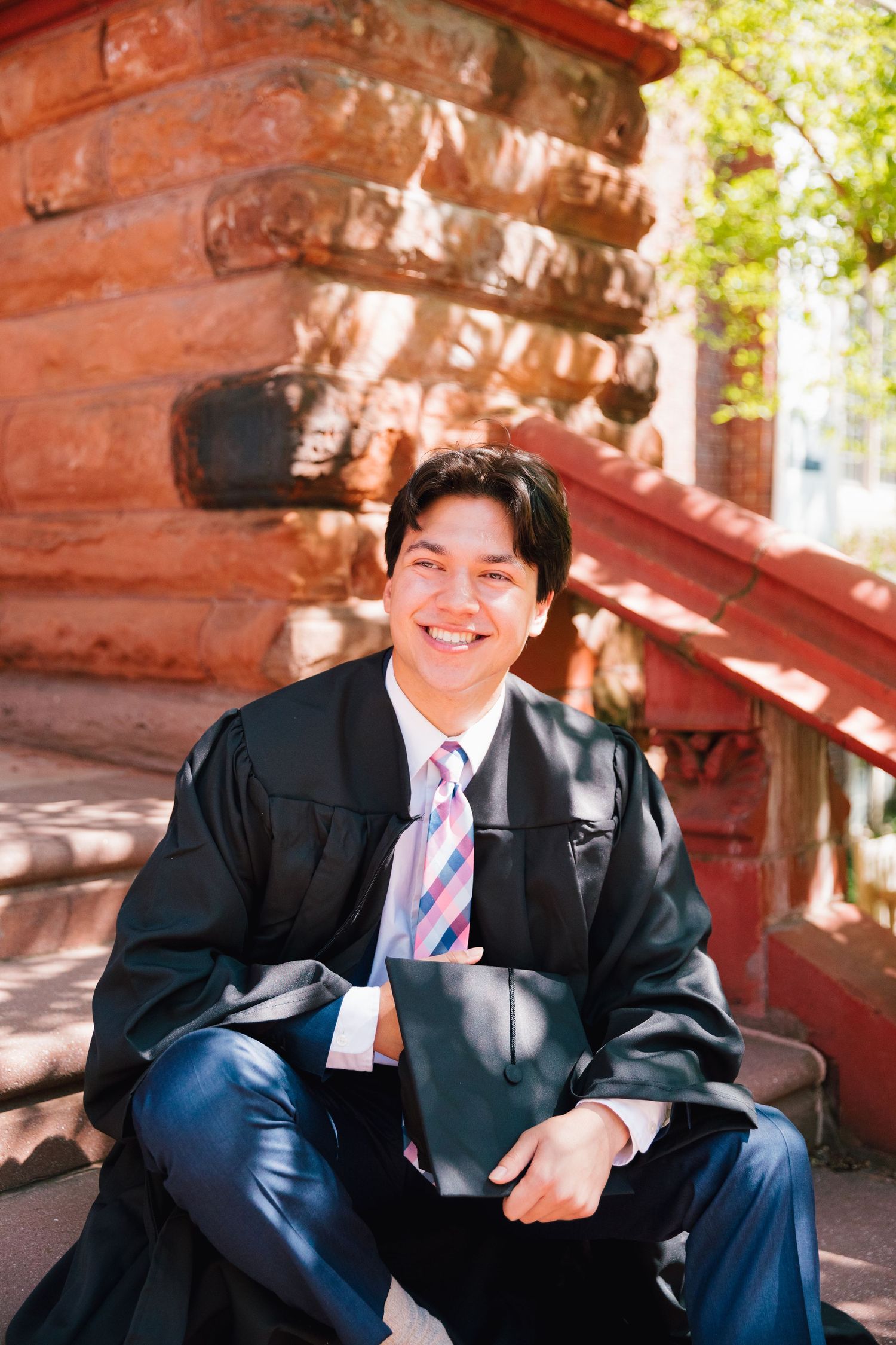 Graduate in formal attire sits casually on red brick stairs during photo session.