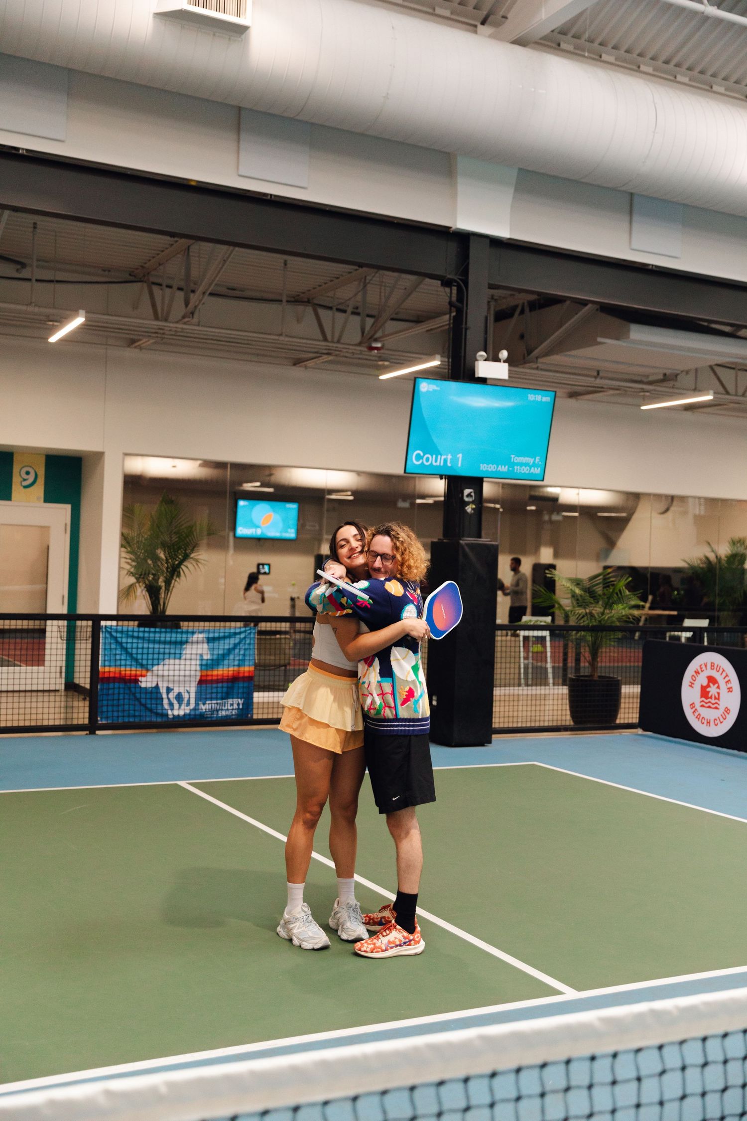 Two players share a warm hug on an indoor pickleball court, holding paddles after their match.