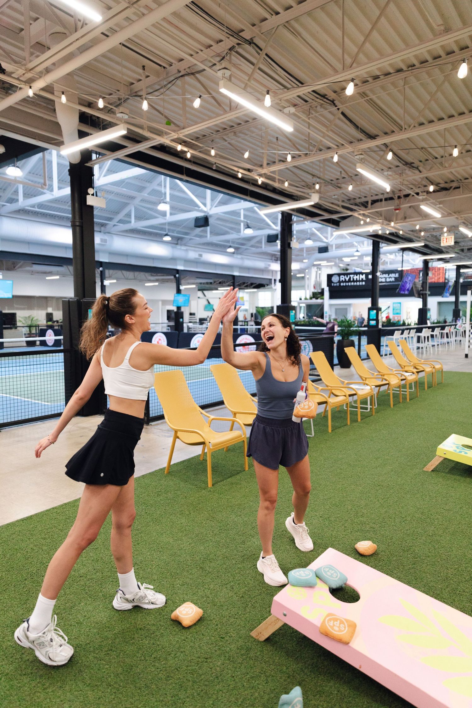 Two women high-fiving while playing cornhole on indoor turf at a modern sports entertainment venue.
