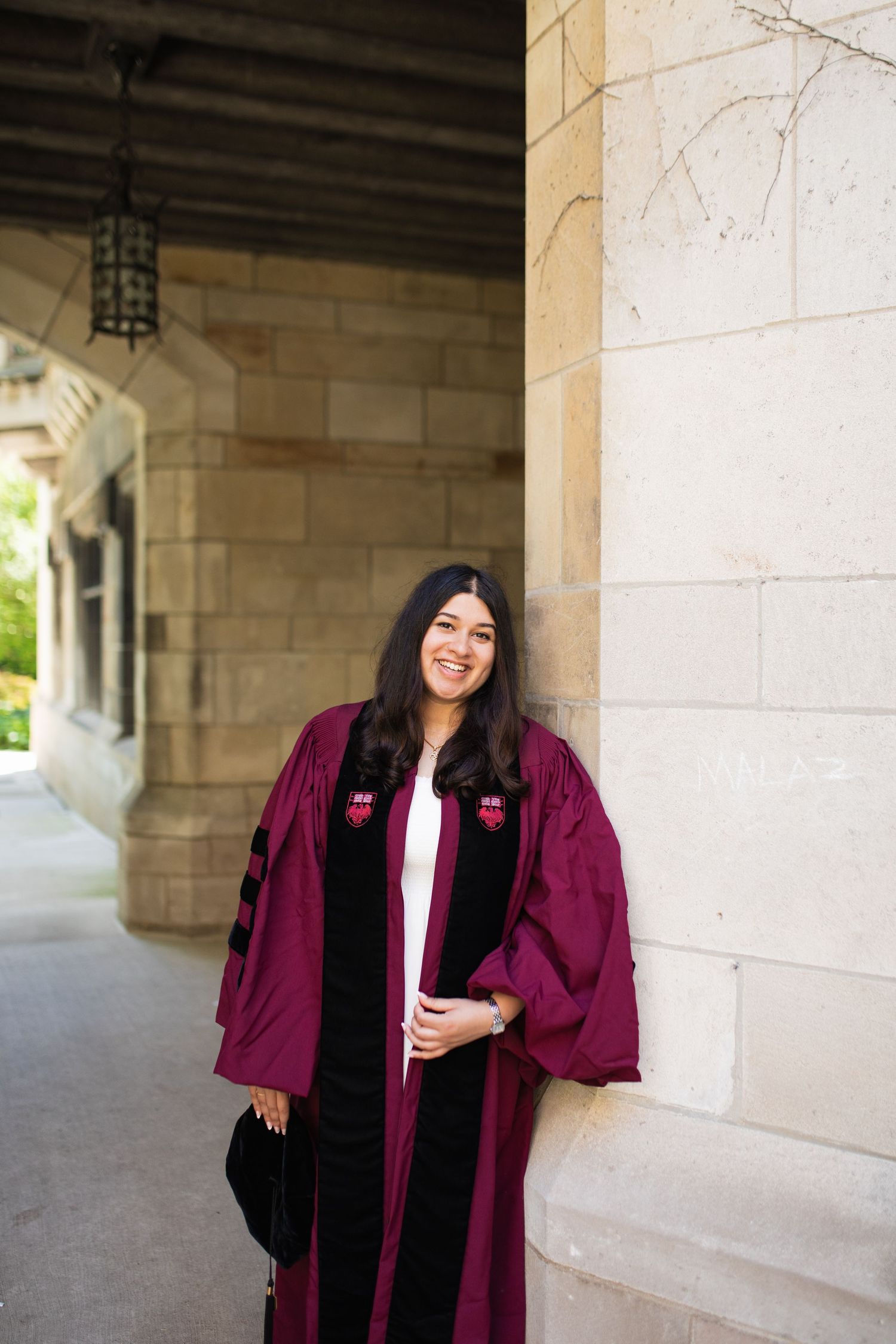 Graduate in maroon academic regalia poses under stone archway.