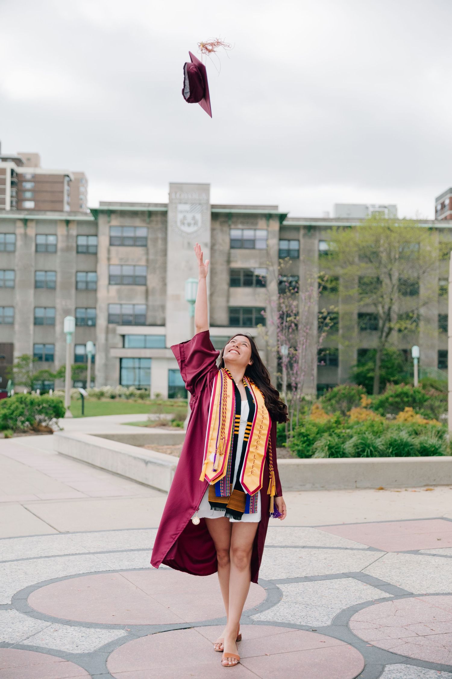 Graduate tosses burgundy graduation cap in air while wearing honors cords on campus.