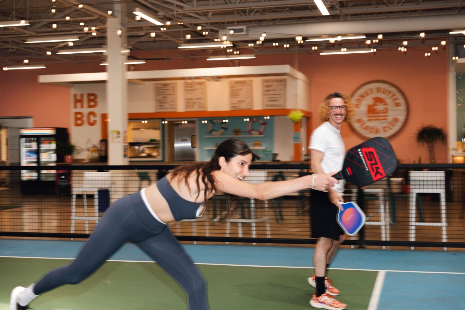 Two players enjoy a pickleball match indoors at a vibrant sports facility with a bright blue court.