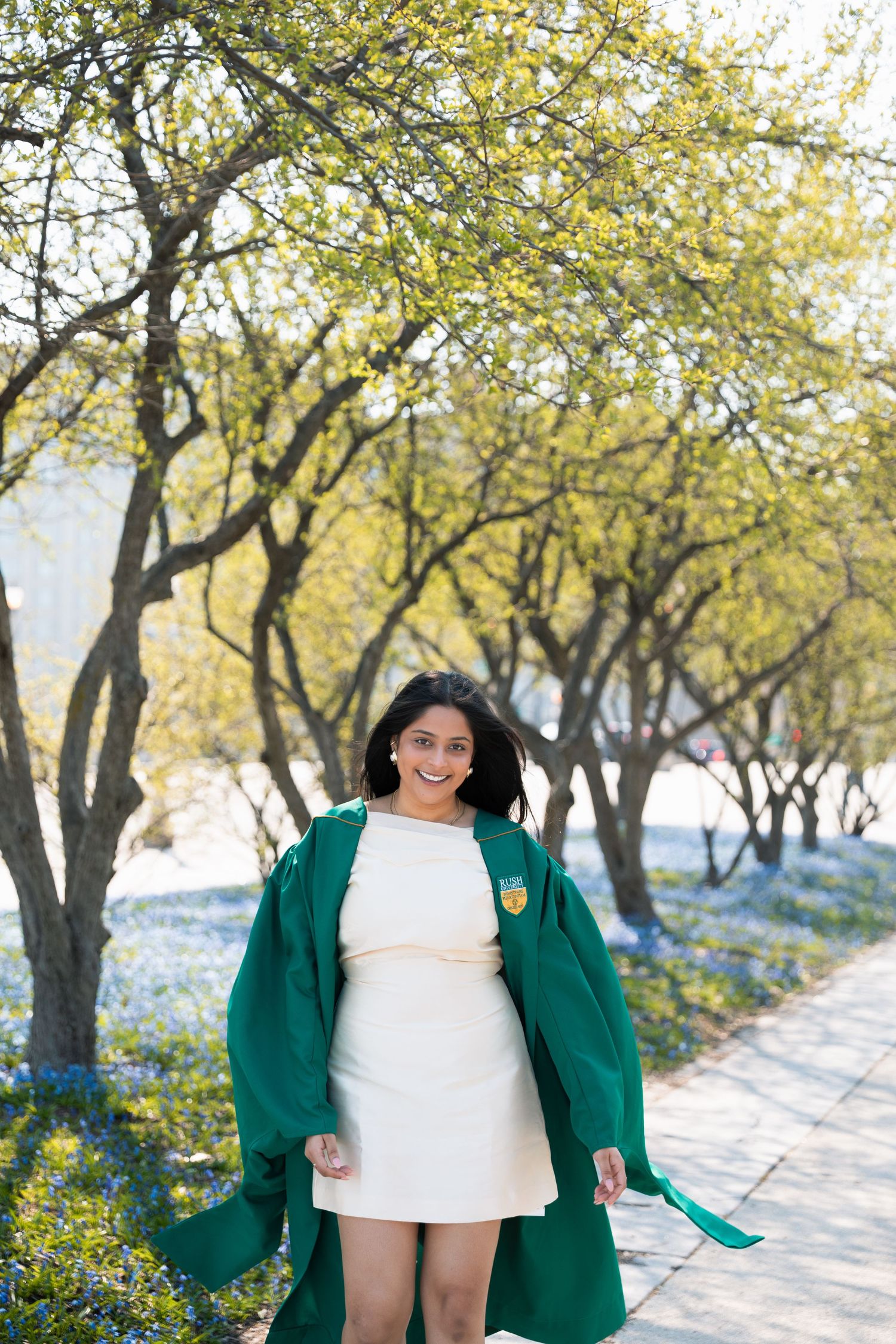 Graduate in green gown walks through path lined with spring trees and blue flowers.