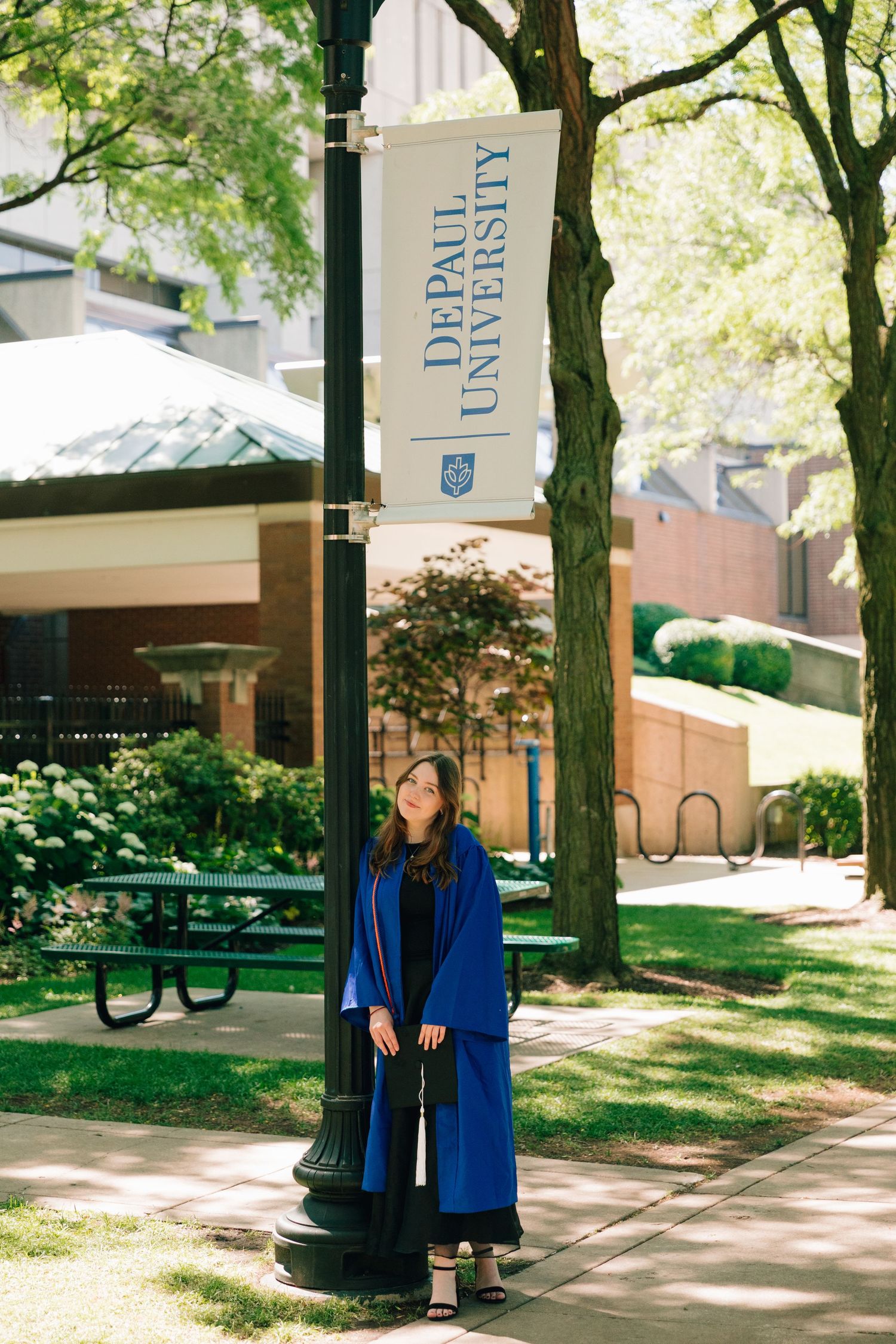 Graduate in blue graduation gown stands beneath university banner on tree-lined campus.