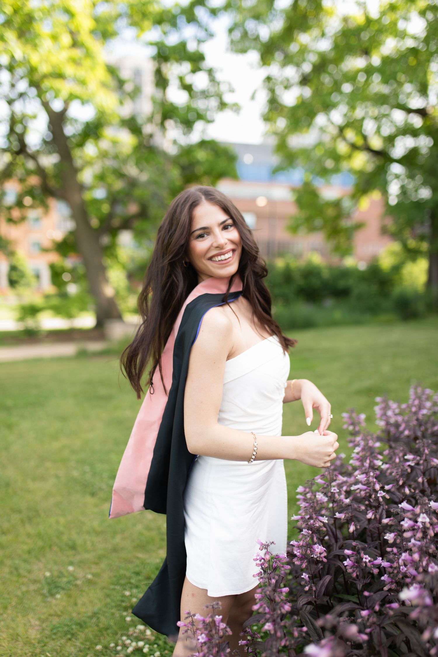 Graduate in white dress poses near blooming purple flowers in garden setting.