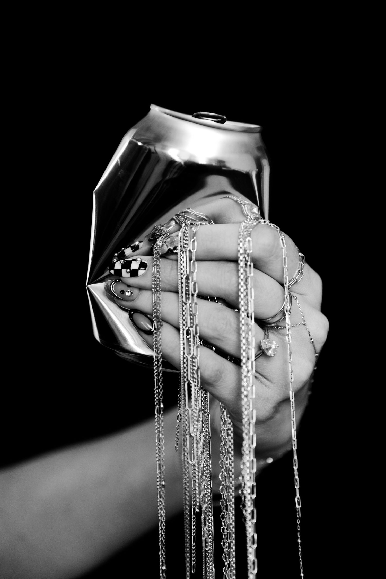 Hand crushing an aluminum can with chains and checkered nail art in dramatic black and white photography.