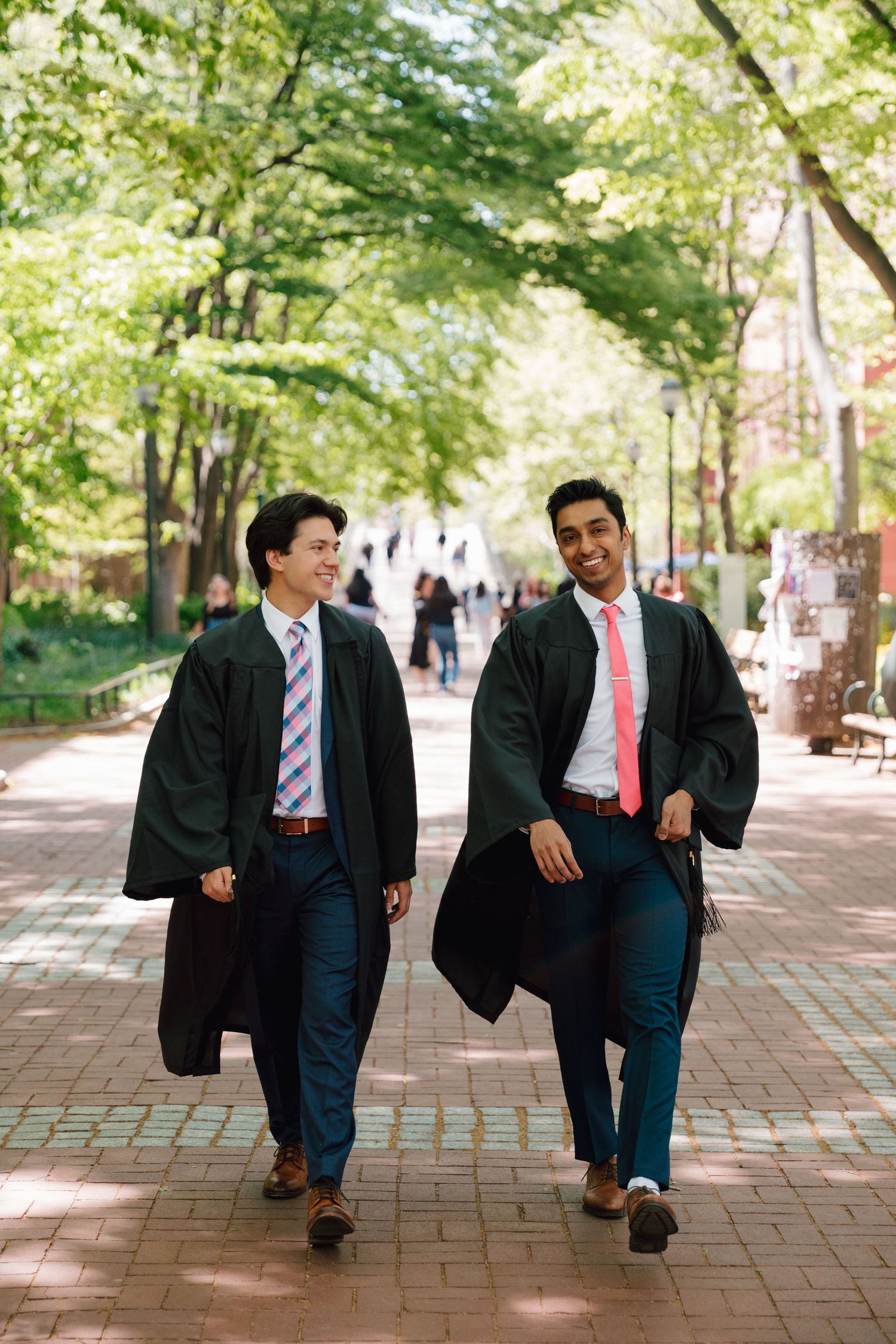 Graduates in suits and ties walk playfully along campus brick pathway.