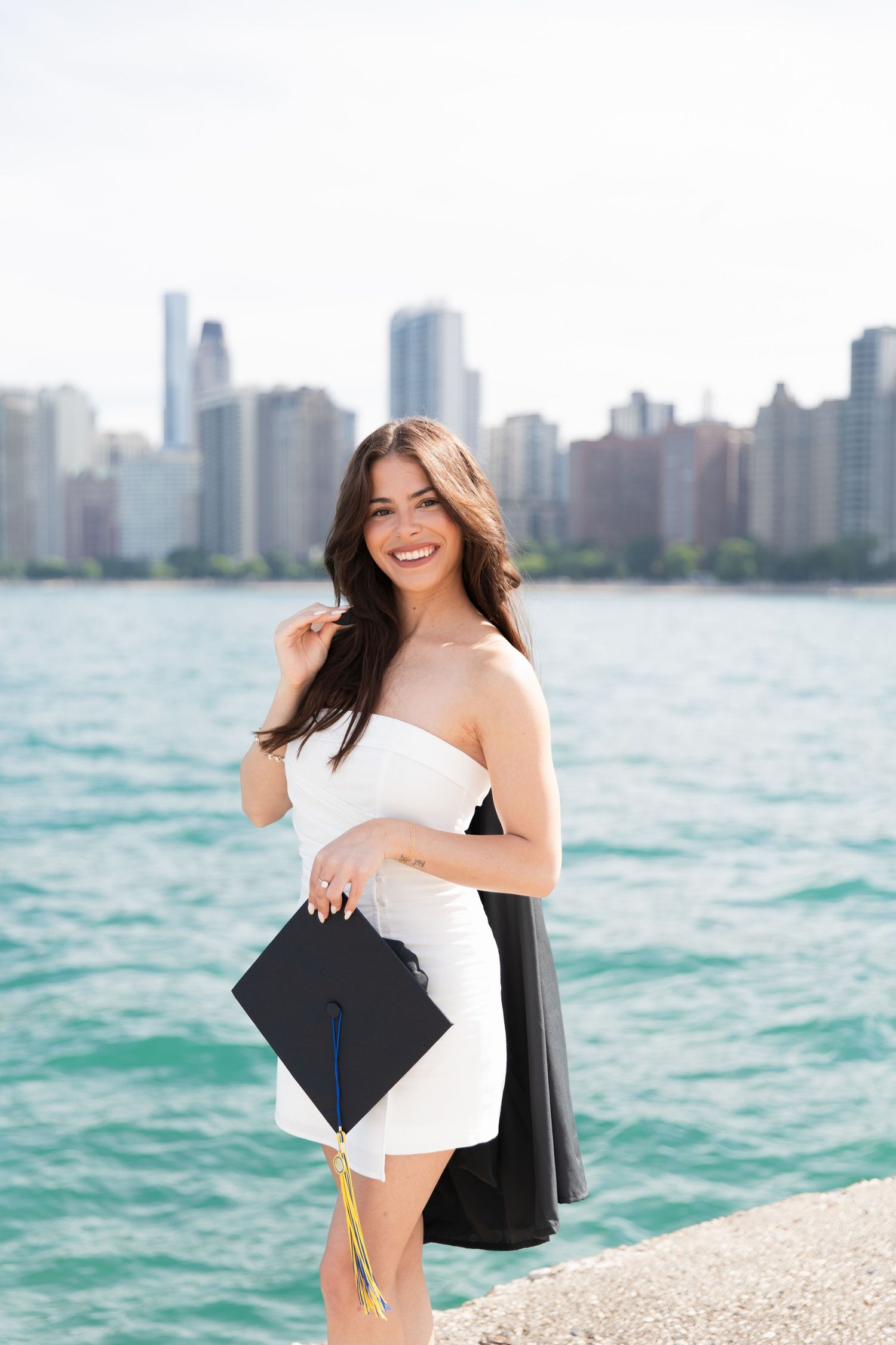 Graduate in white dress holds cap against Chicago skyline and Lake Michigan background.