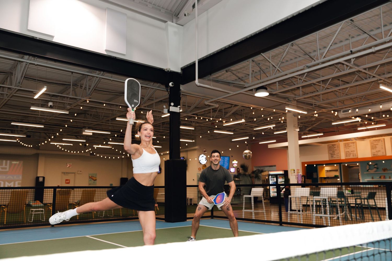 Two players enjoying a pickleball match indoors at a modern recreational facility with court lines visible.