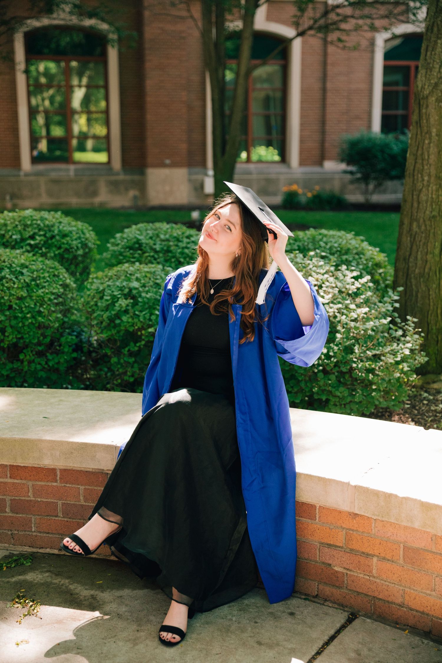 Graduate in blue academic regalia and black dress poses joyfully on campus.