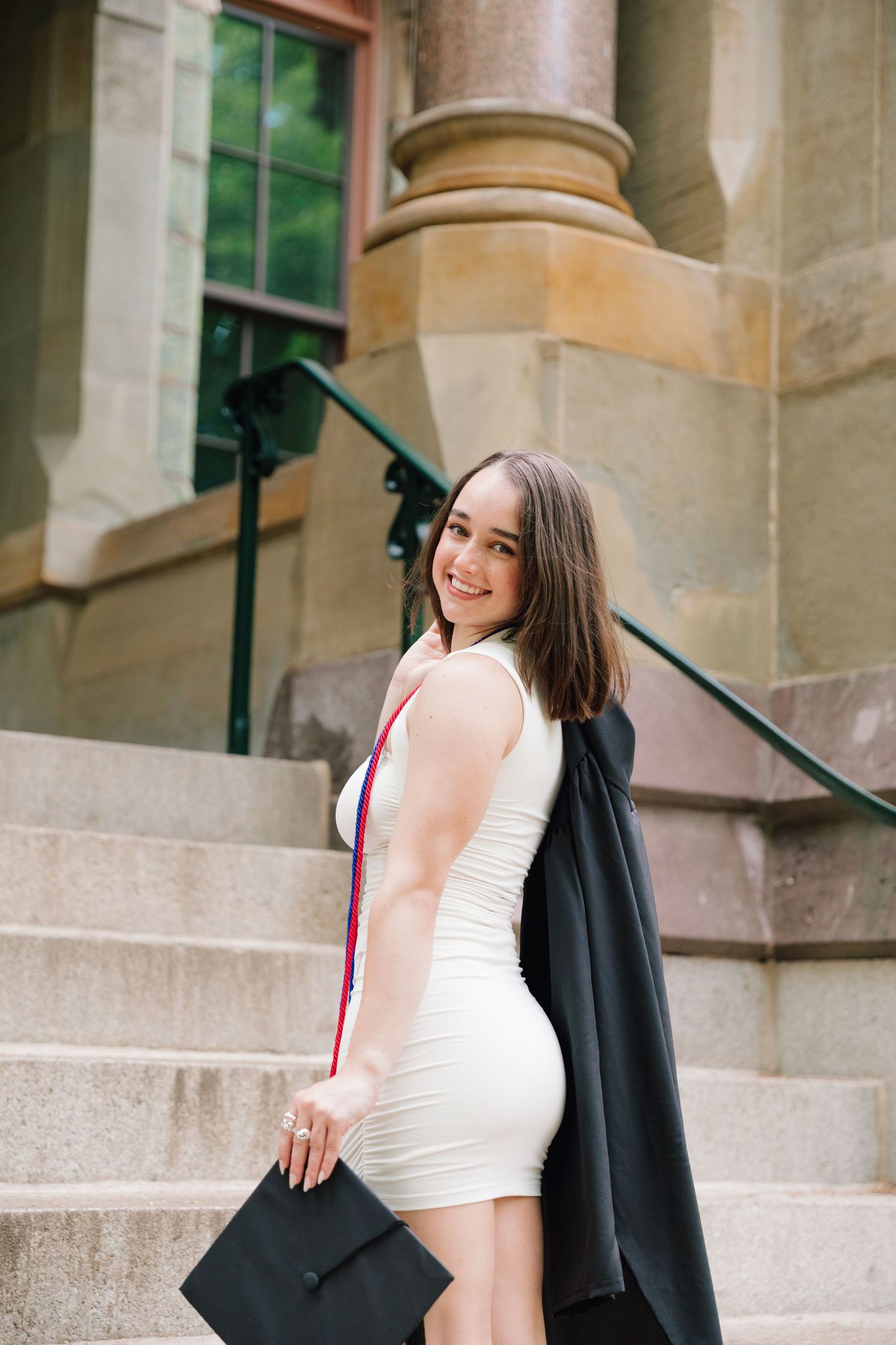 Graduate in white dress and black gown poses on stone steps.