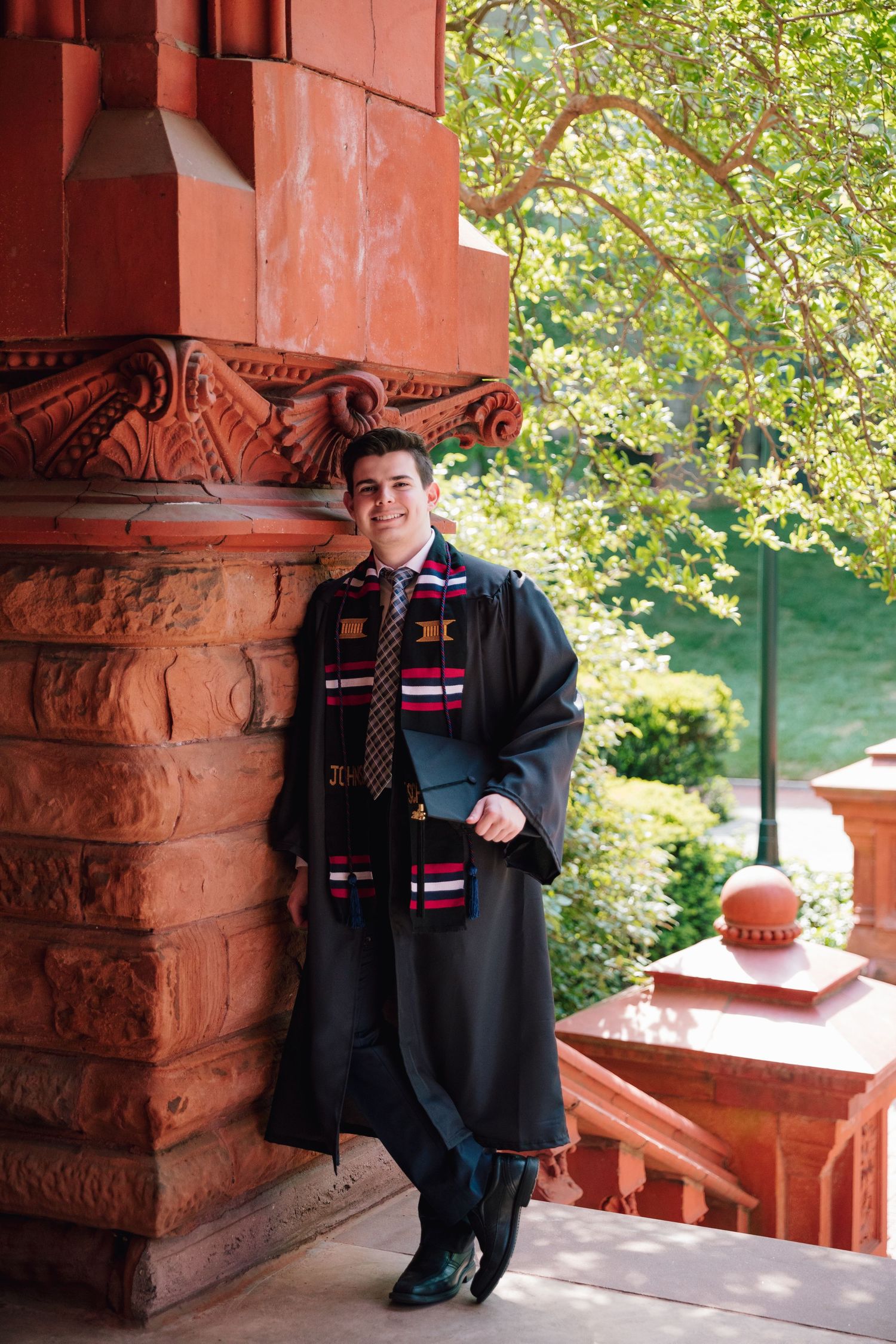 Graduate in academic regalia poses against ornate red stone column on campus.
