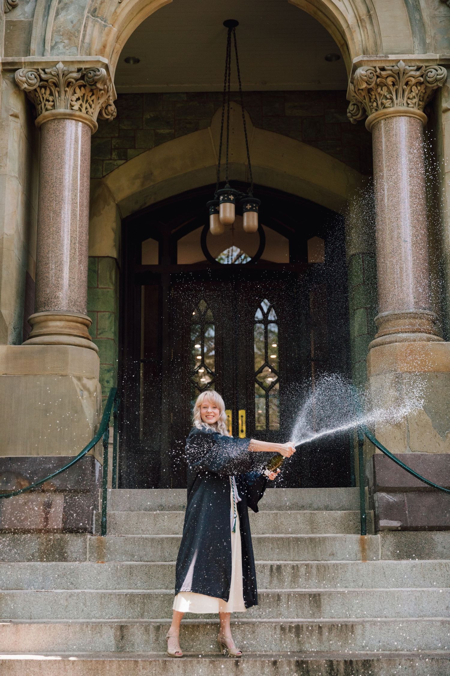 Graduate sprays champagne while celebrating on stone steps of historic building.